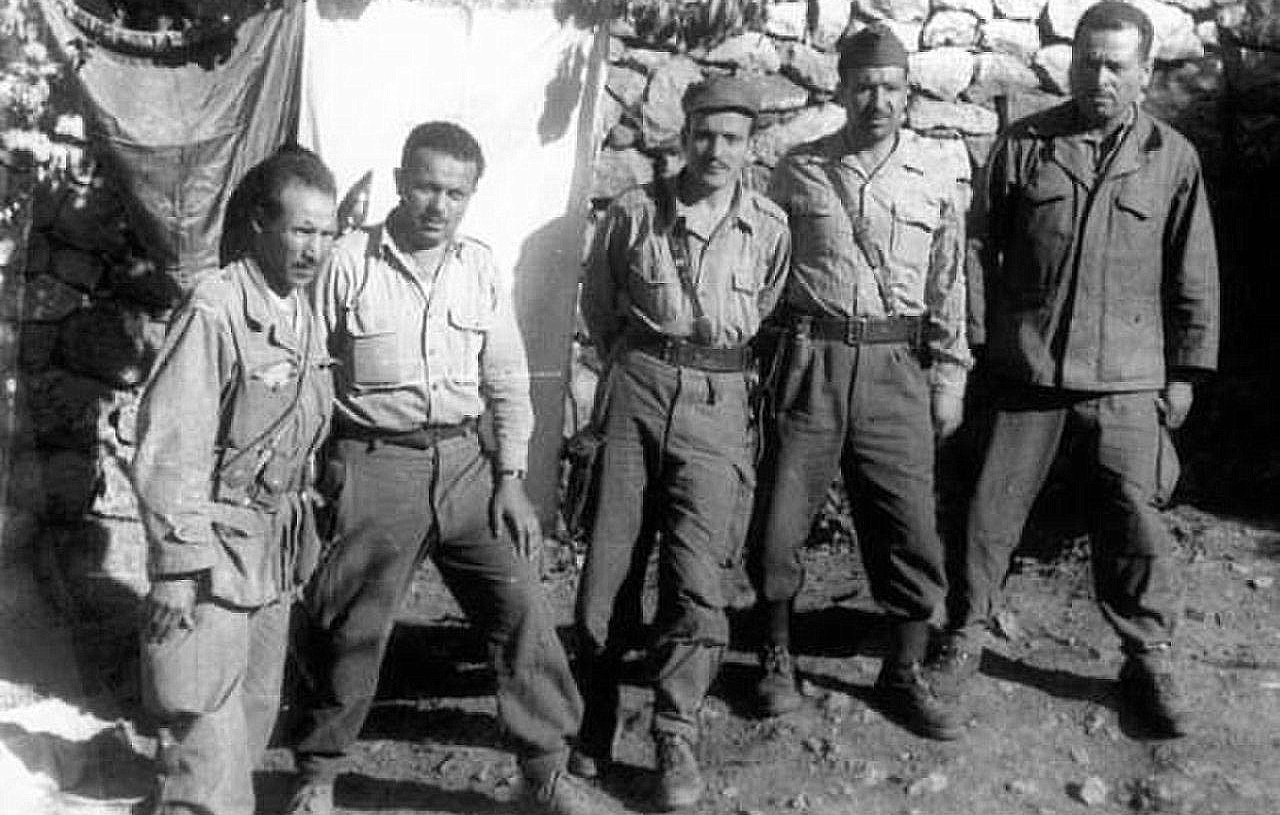 L'image montre un groupe de cinq hommes, probablement des soldats, posant ensemble devant une structure en pierre. Ils portent des uniformes militaires, composés de chemises à manches longues et de pantalons. Les expressions de leurs visages sont sérieuses et déterminées. En arrière-plan, on peut voir une toile suspendue et des pierres qui forment un mur. L'ambiance semble être celle d'un camp militaire.