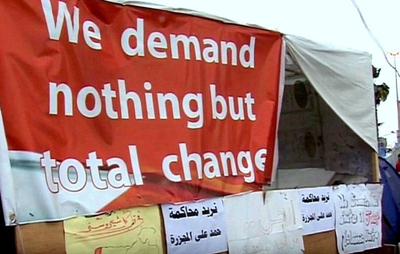 L'image montre une grande pancarte rouge avec le texte "We demand nothing but total change" en anglais, ce qui signifie "Nous exigeons rien d'autre qu'un changement total". En dessous de cette pancarte, on peut voir plusieurs autres affiches, probablement en arabe, qui expriment également des demandes ou des revendications. L'ensemble de la scène évoque un lieu de manifestation ou de protestation, suggérant des aspirations à un changement significatif.