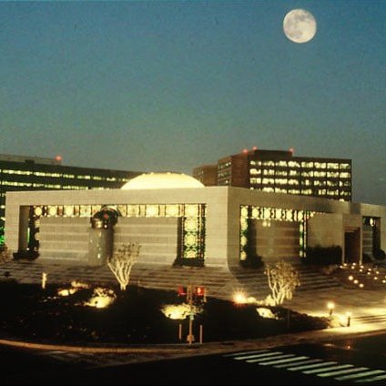 L'image montre un paysage urbain nocturne, avec un bâtiment moderne bien éclairé au centre. Ce bâtiment a une architecture distinctive, avec une coupole et des éléments de design contemporains. À l'arrière-plan, plusieurs autres structures se dressent, leurs fenêtres illuminées créant une ambiance vivante. On peut également voir une grande lune brillante dans le ciel, ajoutant une touche romantique à la scène. Des arbres et des zones paysagées entourent le bâtiment, et des lampadaires éclairent les chemins et les espaces extérieurs, rendant l'ensemble accueillant et esthétique.