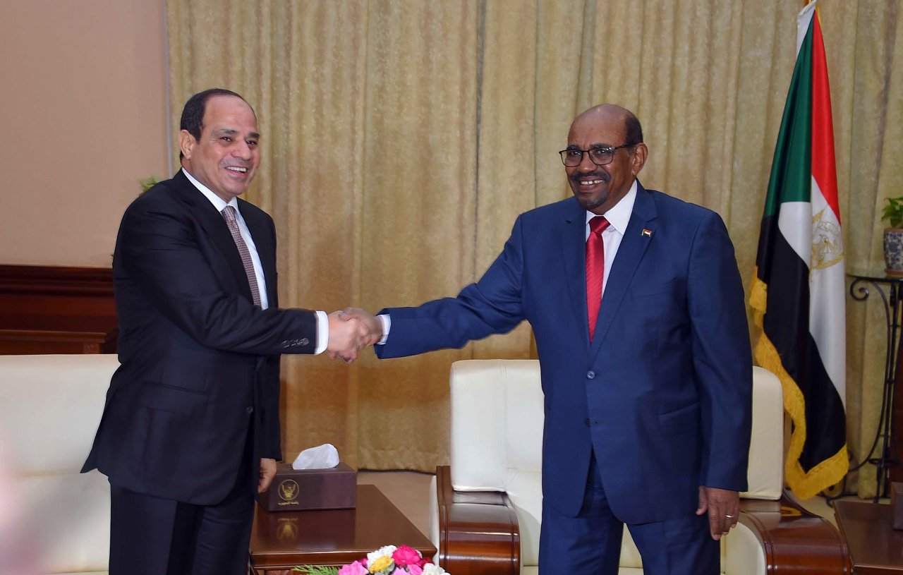 L'image montre deux hommes en train de se serrer la main dans un cadre de rencontre officielle. L'un porte un costume sombre et l'autre un costume bleu. Ils sont entourés d'un décor formel, comprenant des drapeaux et des éléments de décoration. La scène suggère un moment de dialogue ou de coopération entre les deux personnes, probablement des responsables politiques. Un vase avec des fleurs est posé sur la table entre eux.