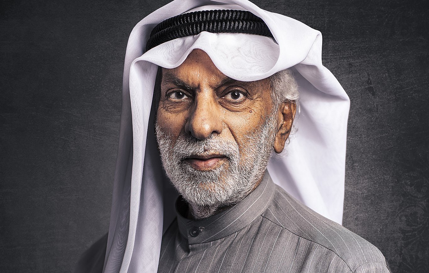 الصورة تظهر رجلاً مسنًا يرتدي الزي التقليدي العربي. لديه لحية رمادية وشعر أبيض، ويرتدي غترة بيضاء مع عقال أسود. يحمل ابتسامة لطيفة ويبدو أنه يتمتع بحضور مهيب. الخلفية مظلمة قليلاً مما يبرز تفاصيل وجهه وملابسه.