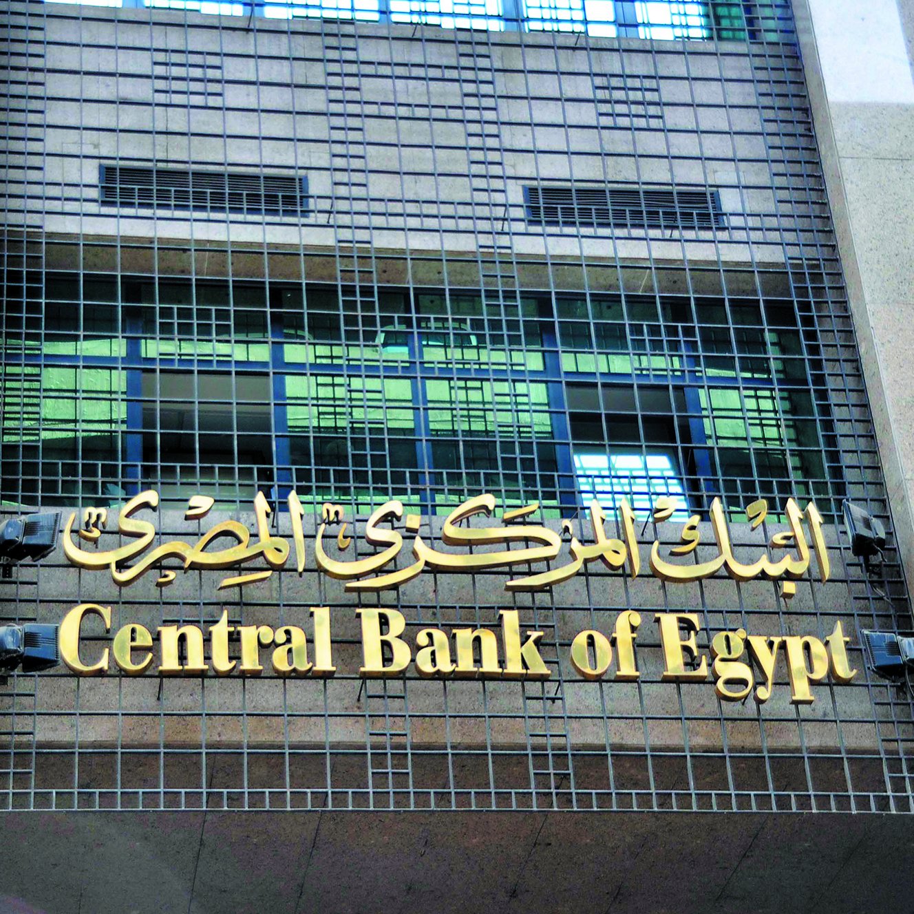 The image features the facade of a building that houses the Central Bank of Egypt. The building has a modern design with a prominent sign displaying "Central Bank of Egypt" in both English and Arabic. The Arabic text is situated above the English, and the overall appearance conveys a sense of official importance and stability. The building is constructed with stone materials, and its facade includes grid-like elements, suggesting a blend of contemporary and traditional architectural styles.