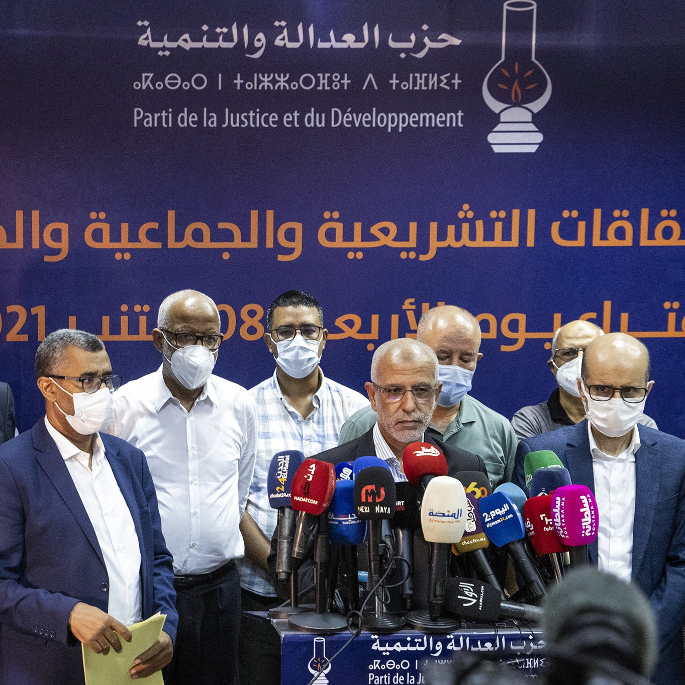 Sur l'image, un groupe d'hommes se tient devant une bannière affichant le nom d'un parti politique, le Parti de la Justice et du Développement. Les hommes portent des masques de protection, ce qui suggère un contexte de santé publique. Ils semblent s'apprêter à faire une déclaration ou une conférence de presse, entourés de microphones et de caméras. L'atmosphère semble sérieuse et professionnelle.