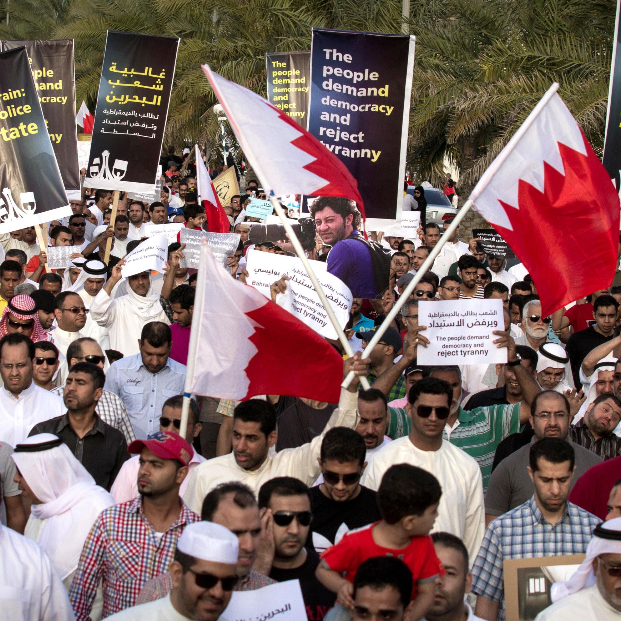 الصورة تظهر تجمعًا حاشدًا لمتظاهرين في البحرين يحملون لافتات وأعلام البلاد. يتواجد الأشخاص في مكان مفتوح، ويسود الجو شعور بالتضامن والمطالبة بالتغيير. اللافتات تتضمن مطالب للديمقراطية ورفض السياسات القمعية. يرتدي المتظاهرون ملابس تقليدية وطنية، ويبدو أن الحشد متنوع من حيث الأعمار.