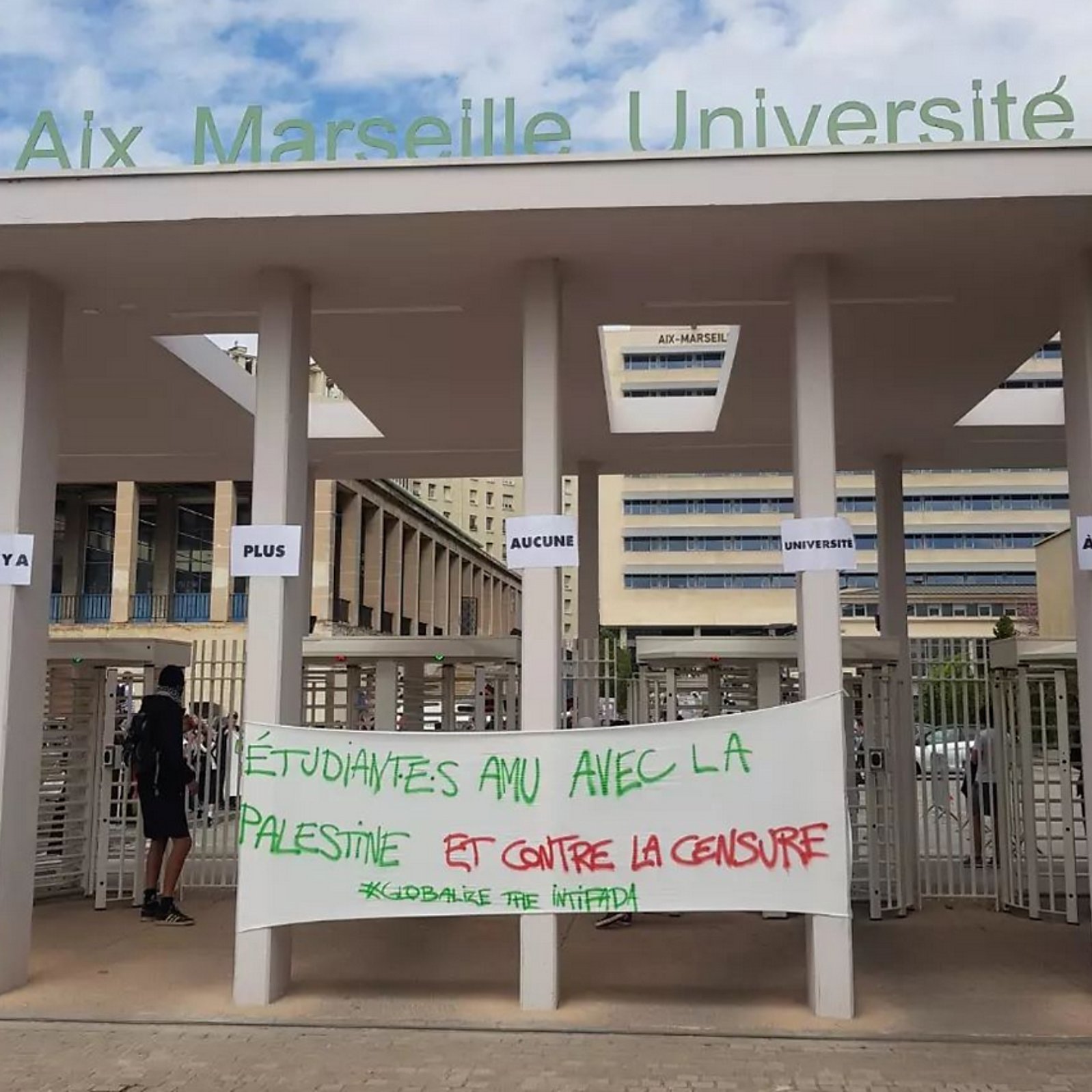 L'image montre l'entrée d'Aix Marseille Université, avec des panneaux affichant des messages. Au premier plan, il y a une grande bannière sur laquelle est écrit en rouge et en vert : "Étudiantes AMU avec la Palestine et contre la censure". L'atmosphère semble refléter un soutien à la cause palestinienne et une dénonciation de la censure. Quelques personnes sont visibles dans la scène, et le cadre est universitaire.