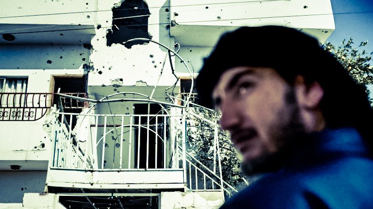 L'image montre un bâtiment portant des marques visibles d'impacts de balles, suggérant des dommages dus à des conflits. Au premier plan, un homme est partiellement visible, tourné vers l'extérieur, avec une expression pensive. L'environnement semble urbain, et le contraste entre le visage de l'homme et le bâtiment endommagé crée une atmosphère de tension et de tristesse.