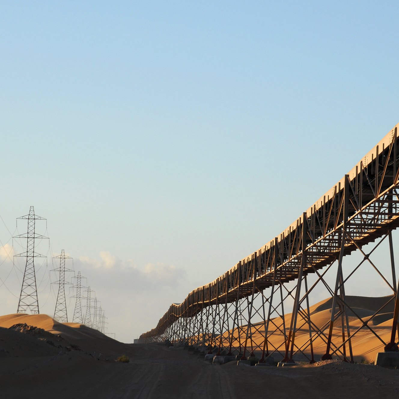 L'image montre un long convoyeur à bande surélevé qui s'étend à travers un paysage désertique. Les dunes de sable se dressent en arrière-plan, illuminées par la lumière dorée du soleil, créant un contraste frappant avec la structure métallique du convoyeur. De chaque côté de la route, des pylônes électriques s'élèvent, ajoutant une dimension industrielle à la scène. Le ciel est clair, suggérant une atmosphère paisible malgré la vaste étendue désertique.