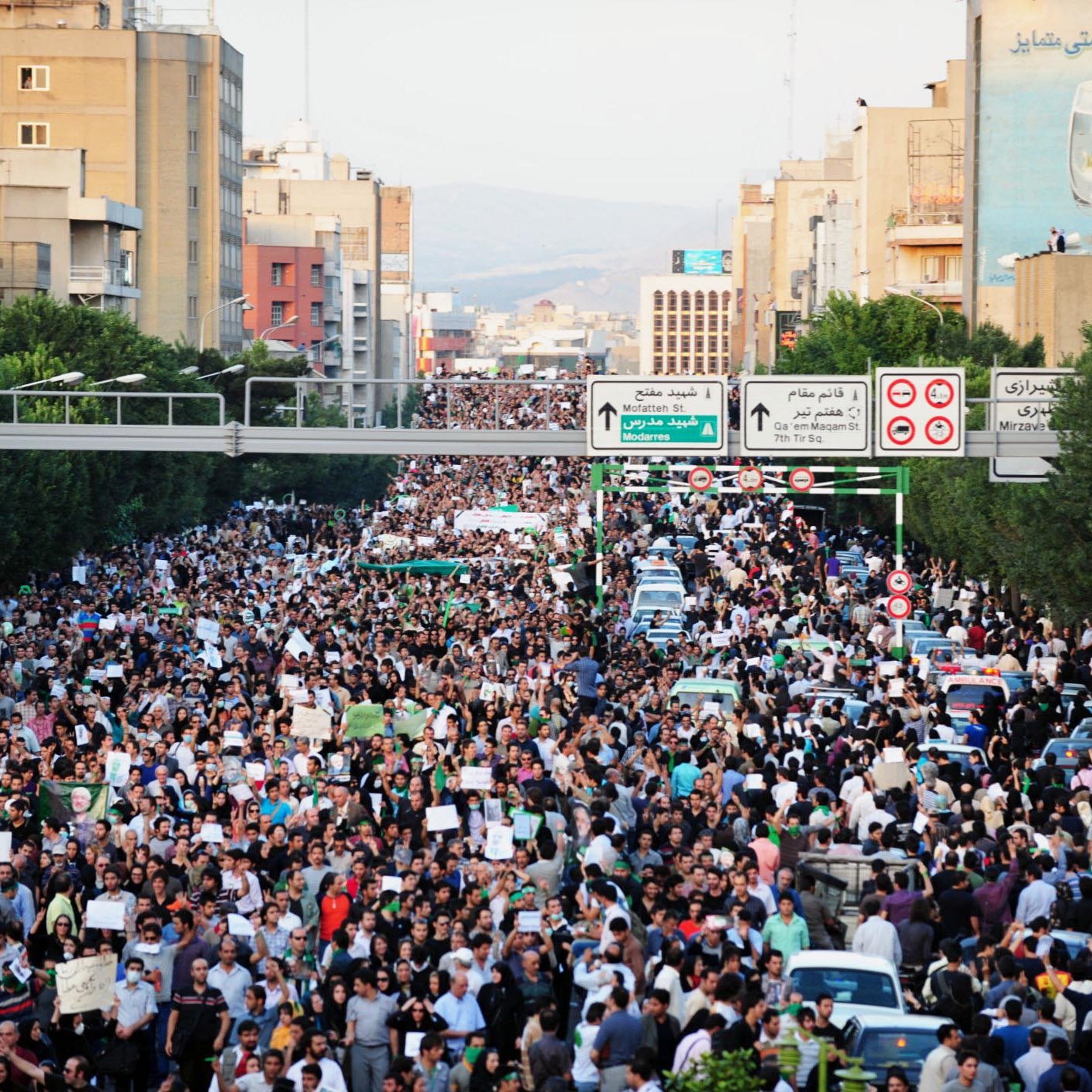 L'immagine mostra una grande folla di manifestanti in una strada cittadina. Le persone sembrano riunite per un evento significativo, con molti che portano cartelli e striscioni. I palazzi al bordo della strada sono visibili, così come alcune automobili parcheggiate. La scena trasmette una forte sensazione di unione e mobilitazione collettiva. Le strade sono affollate e l'atmosfera è intensa, suggerendo che si tratta di una protesta o di una manifestazione pubblica.