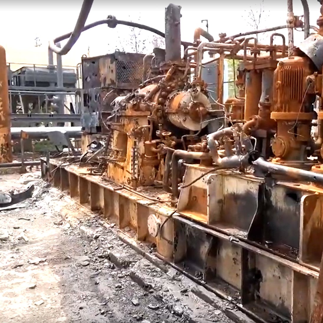 L'image montre une installation industrielle endommagée, probablement suite à un incendie. On peut voir des équipements métalliques, tels que des machines et des tuyaux, recouverts de rouille et de suie. Le sol est noirci par des résidus, indiquant les conséquences d'un sinistre. L'environnement semble très dégradé, avec des éléments de construction en arrière-plan également affectés par des dommages. L'ensemble donne une impression de désolation et de négligence.
