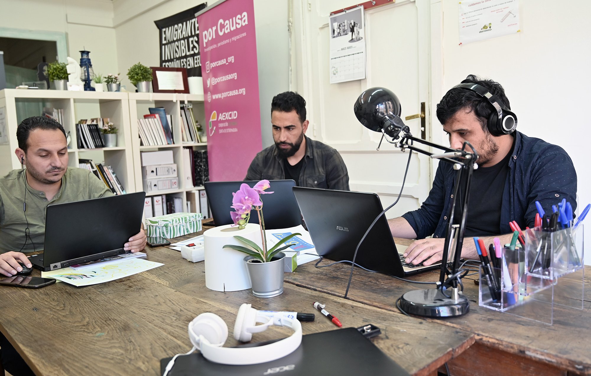 L'image montre trois hommes assis autour d'une table en bois, chacun travaillant sur un ordinateur portable. Ils semblent concentrés sur leurs tâches. Sur la table, on peut voir des objets tels qu'un casque, des stylos et une plante en pot. À l'arrière-plan, il y a des affiches et des livres, ce qui donne une ambiance de bureau ou d'espace de travail collaboratif. Un microphone est également visible sur la table, ce qui pourrait indiquer qu'ils sont en train d'enregistrer ou de diffuser quelque chose.