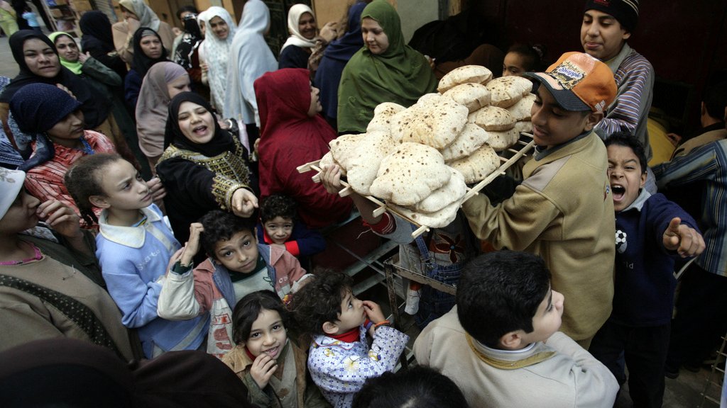 L'image montre une scène animée dans une rue, probablement dans un pays arabe. Un jeune garçon porte un plateau chargé de pains, entouré d'une foule de personnes, comprenant des adultes et des enfants. On peut voir des visages souriants et des expressions variées, ce qui donne une impression de convivialité et d'effervescence. Les gens semblent attendre pour acheter du pain, et la diversité des vêtements suggère une ambiance culturelle riche. L'image capte un moment de vie quotidienne, centré sur l'échange et la nourriture.
