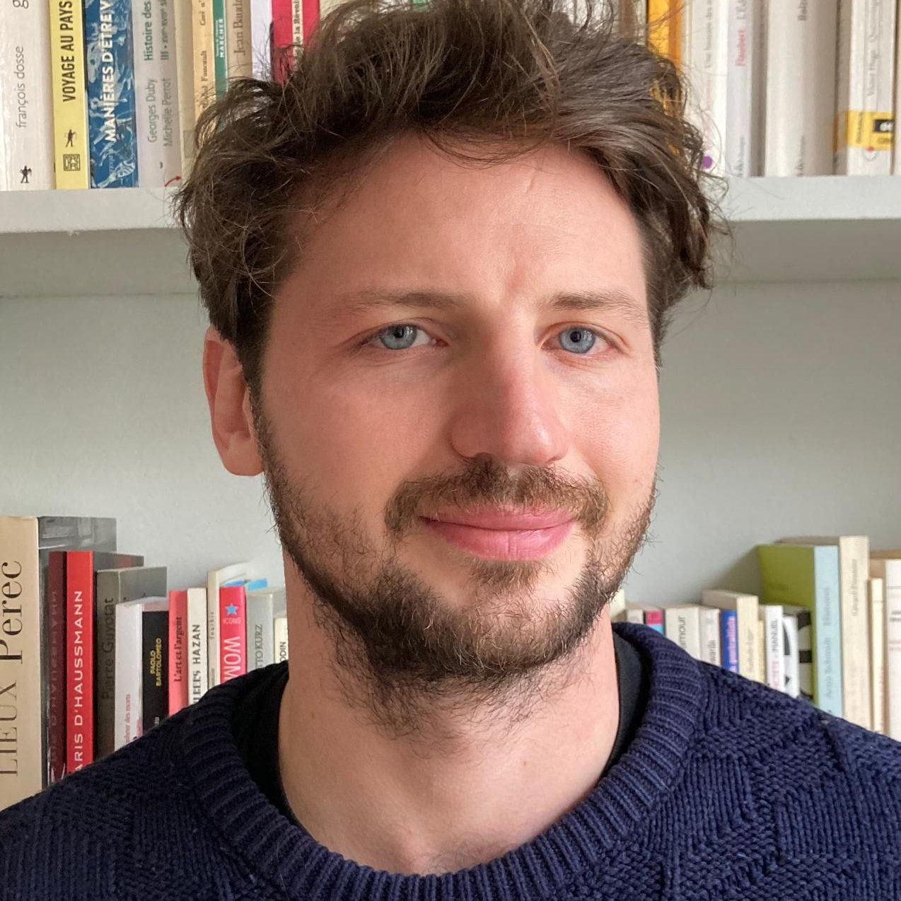Homme souriant avec des cheveux bruns et une barbe, devant une étagère de livres.