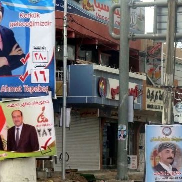 L'image montre une rue animée bordée de panneaux électoraux. On peut y voir plusieurs affiches de campagne, chacune présentant différents candidats avec leurs noms et numéros associés, ainsi que des logos de partis politiques. Les couleurs des affiches varient, attirant l'attention des passants. L'environnement urbain semble typique d'une zone commerçante, avec des boutiques et des enseignes visibles en arrière-plan.