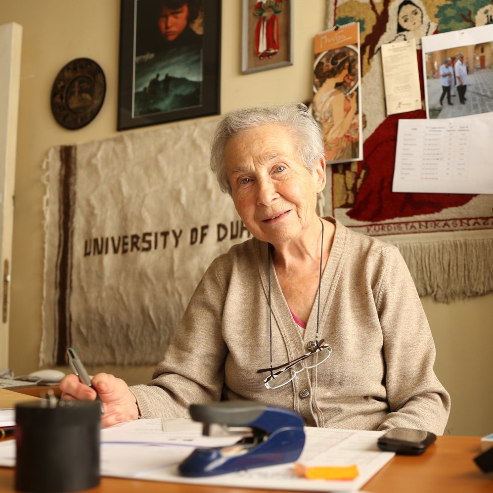 الصورة تظهر امرأة مسنّة تجلس أمام مكتب مغطى ببعض الأوراق والأدوات المكتبية مثل مشبك الورق. يبدو عليها الودّ والسعادة، حيث تبتسم وتضع نظاراتها على ملصق مرتخي حول رقبتها. خلفها، يمكن رؤية جدران مزينة ببعض الصور والإطارات، وكذلك قطعة قماش تحمل اسم "جامعة دبي". أجواء الصورة تعكس بيئة عمل ومتعة في التواصل والذكريات.