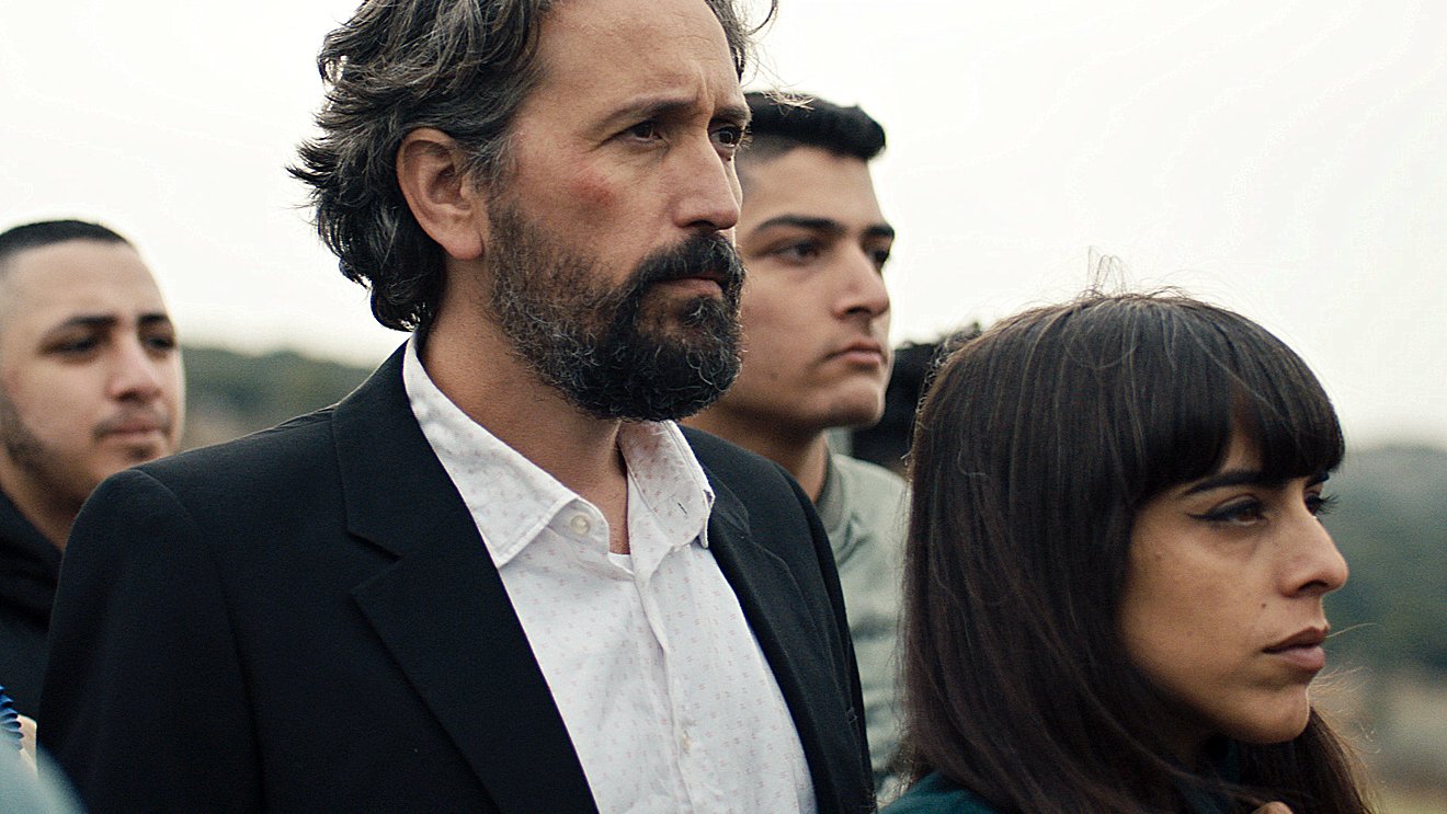 L'image montre un groupe de personnes rassemblées, avec un homme au premier plan, portant un costume noir et une chemise blanche. Il a les cheveux gris et une barbe. À côté de lui se trouve une femme avec des cheveux longs et bruns, qui semble pensive. En arrière-plan, d'autres personnes sont présentes, affichant des expressions sérieuses. L'ambiance général semble solennelle.