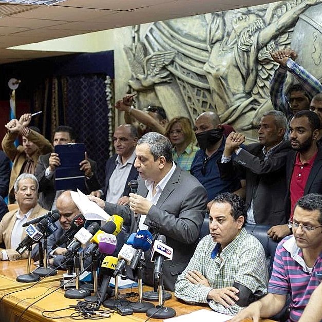 L'image montre une conférence de presse ou une réunion. Un homme se tient au centre, parlant devant un groupe de journalistes, avec plusieurs microphones devant lui. Derrière lui, des personnes semblent exprimer leur soutien ou leur désaccord, levant les poings. L'arrière-plan présente une sculpture en relief qui ajoute de la profondeur à la scène. L'atmosphère semble tendue et engagée.
