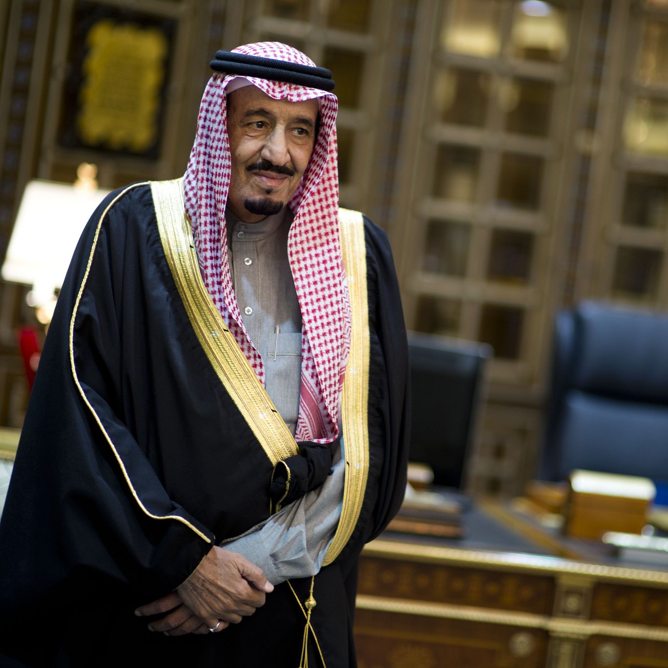 L'image montre un homme en tenue traditionnelle saoudienne, portant une robe noire ornée d'or et un keffieh, une coiffe typique. Il se tient dans un bureau richement décoré, avec des meubles en bois et un éclairage sophistiqué. L'ambiance est formelle et élégante.