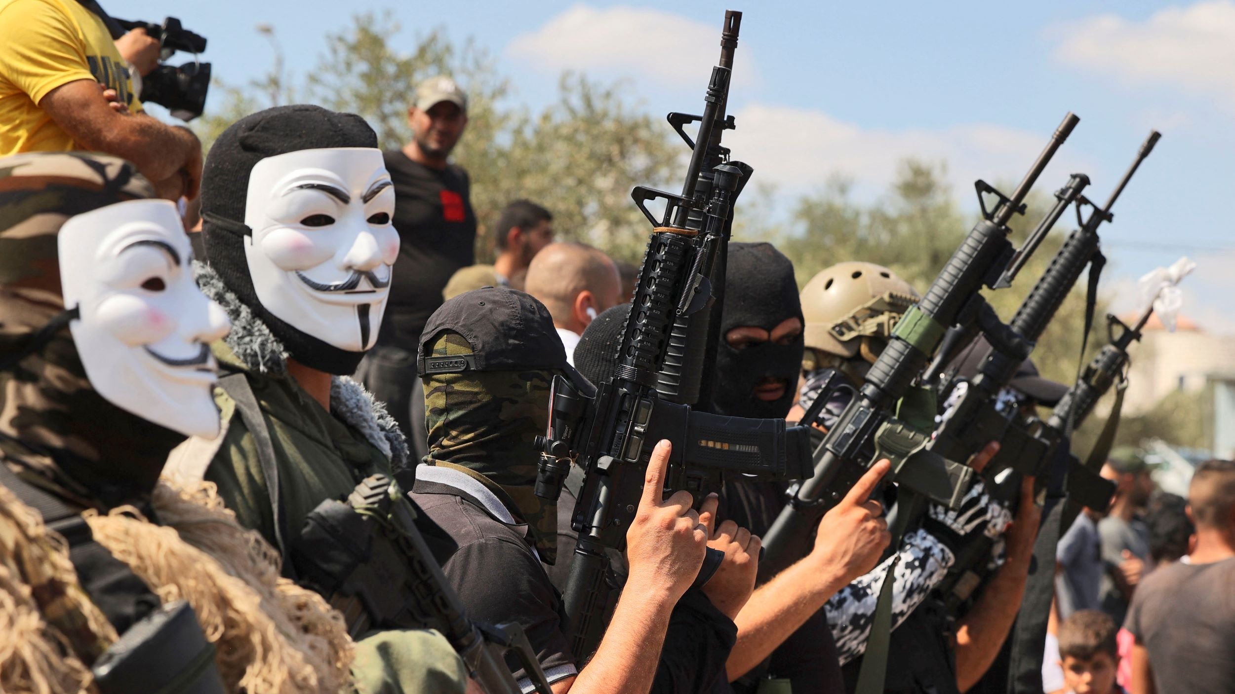 در این تصویر، گروهی از مردان با ماسک‌های سفید و مشکی که صورت‌هایشان را می‌پوشاندند، در حال ایستادن هستند. آن‌ها سلاح‌های اتوماتیک در دست دارند و لباس‌های نظامی به تن دارند. در پس‌زمینه، افراد دیگری به وضوح دیده می‌شوند که به این گروه نگاه می‌کنند. فضا به نظر حالت نظامی و جدی دارد.