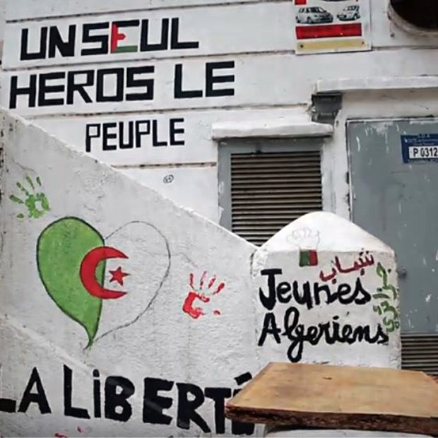 L'image montre un mur peint avec plusieurs graffitis. On y trouve des mots et des phrases exprimant des messages politiques et sociaux. Un dessin représentant un cœur dans les couleurs du drapeau algérien (rouge et vert) est visible. Il y a aussi une inscription en arabe, ainsi que des mots tels que "Jeunies Algériens" et "LA LIBERTÉ". Le fond suggère un lieu qui pourrait être un espace public, témoignant d'une expression artistique et militante.