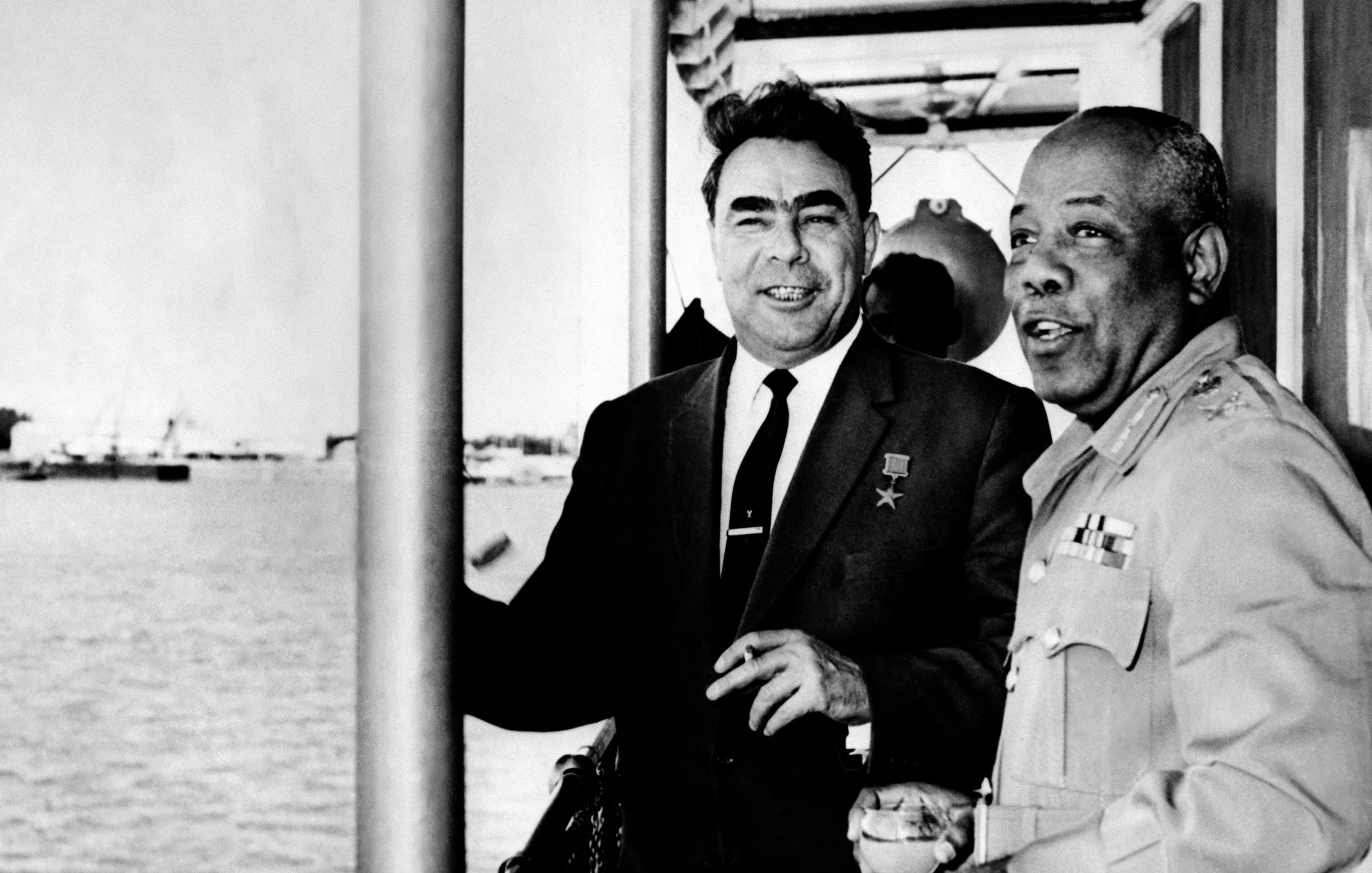 L'image montre deux hommes en train de discuter sur ce qui semble être un bateau ou une plateforme près de l'eau. L'un des hommes porte une médaille, indiquant honneur ou reconnaissance, et les deux semblent souriants et détendus. En arrière-plan, on peut voir des bateaux et une vue maritime. L'atmosphère générale de l'image semble amicale et conviviale.