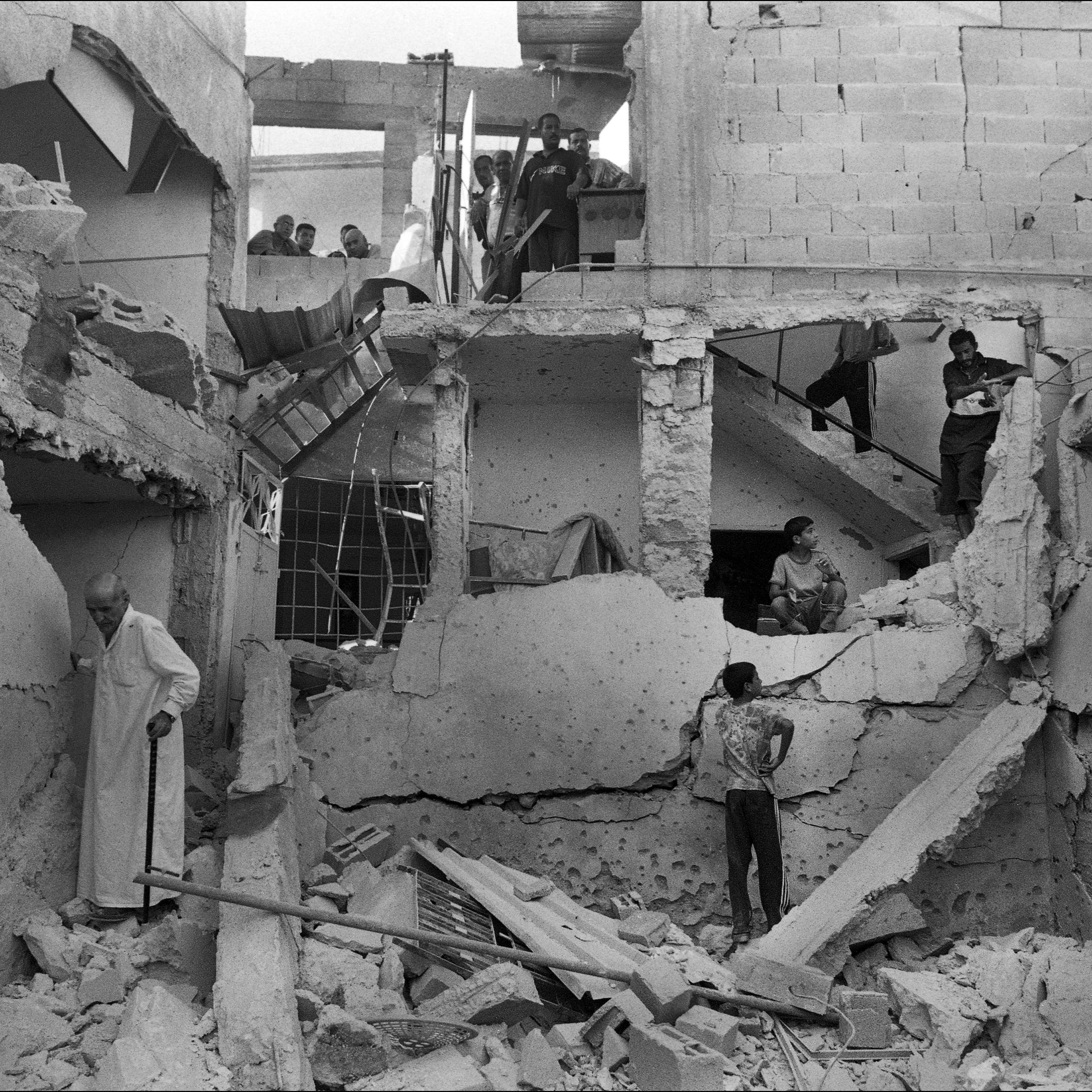 L'image montre un bâtiment en ruines, visiblement endommagé par un conflit ou un désastre. Les murs sont effondrés et des débris jonchent le sol. Plusieurs personnes se trouvent dans l'espace, certains semblent fouiller parmi les débris, tandis que d'autres observent la scène depuis un étage supérieur. L'ambiance est sombre et témoigne des conséquences dévastatrices sur la vie humaine et les infrastructures.