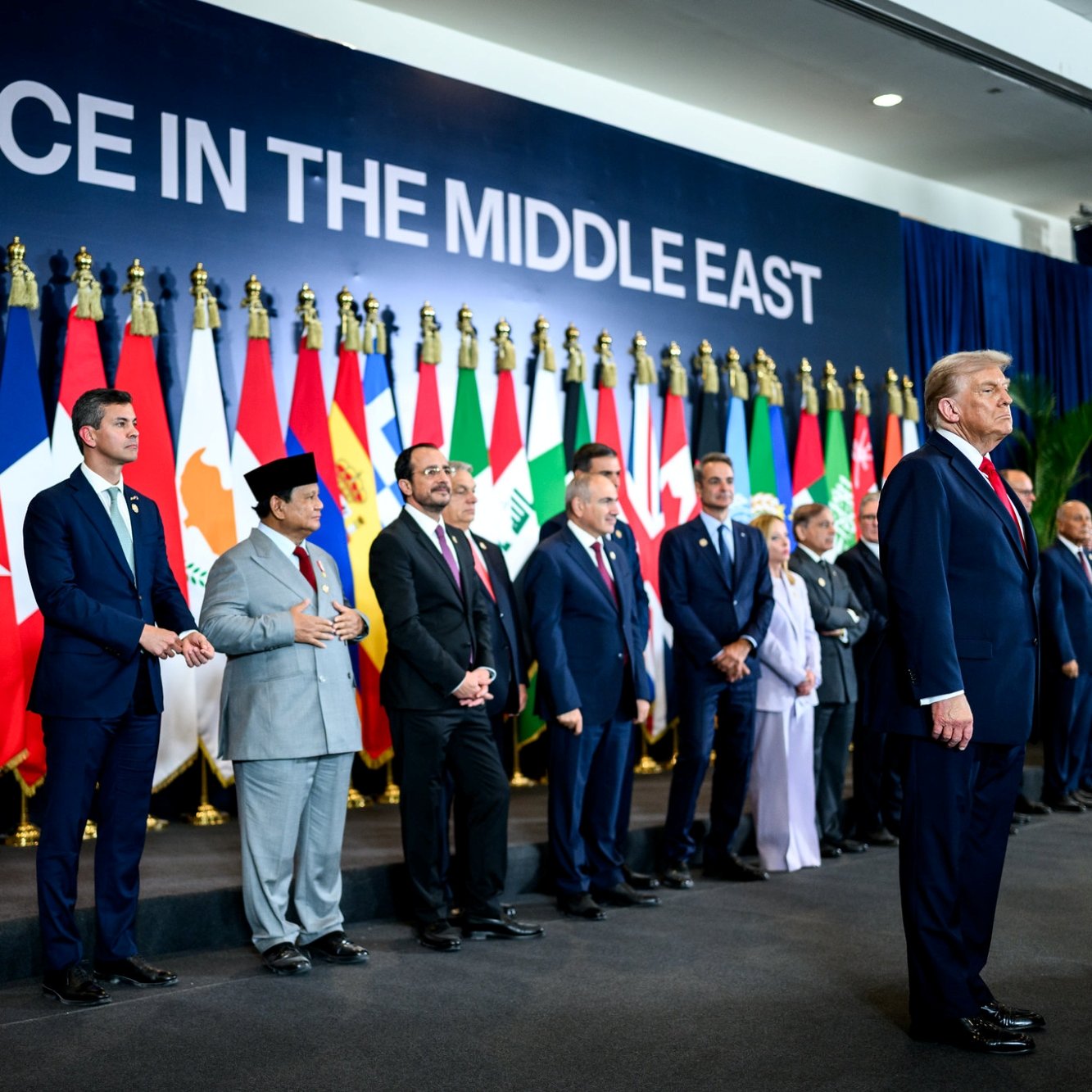 مردان در یک صف ایستاده‌اند، پرچم‌های کشورهای مختلف پشت سرشان و نوشته‌ای بزرگ درباره "صلح در خاورمیانه" بالای سر.