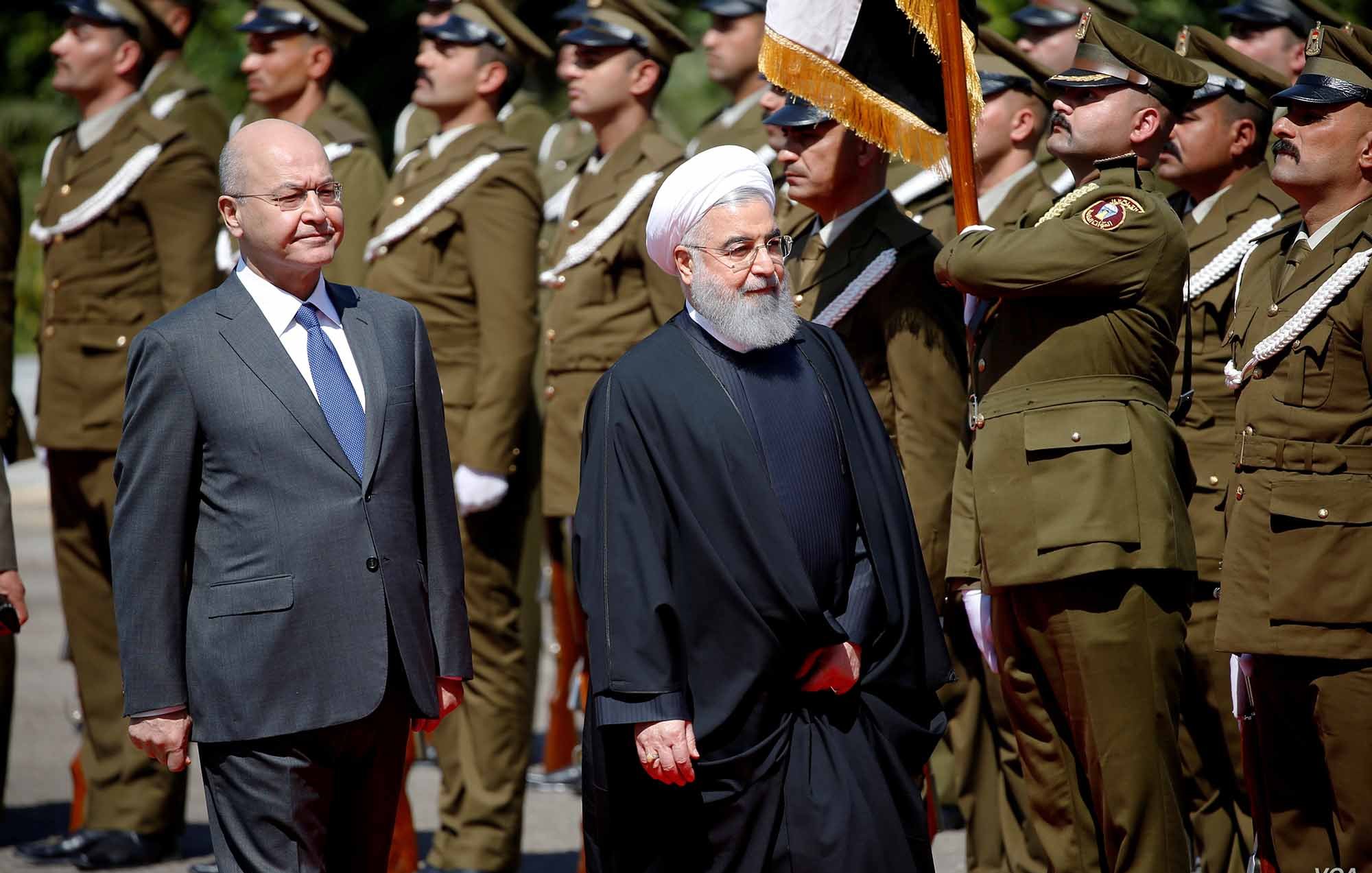 الصورة تظهر شخصيتين سياسيين يسيران معًا، حيث يبدو أنهما في مراسم رسمية. الشخص الأول يرتدي بدلة رسمية، بينما الشخص الثاني يرتدي زيًا تقليديًا أسود مع عمامة. خلفهم يظهر حرس الشرف يرتدون uniforms عسكرية، ويحملون الأعلام. الأجواء تعكس طابعًا رسميًا وجديًا، كما أن الإضاءة واضحة مما يدل على أنها تم التقاطها خلال النهار.