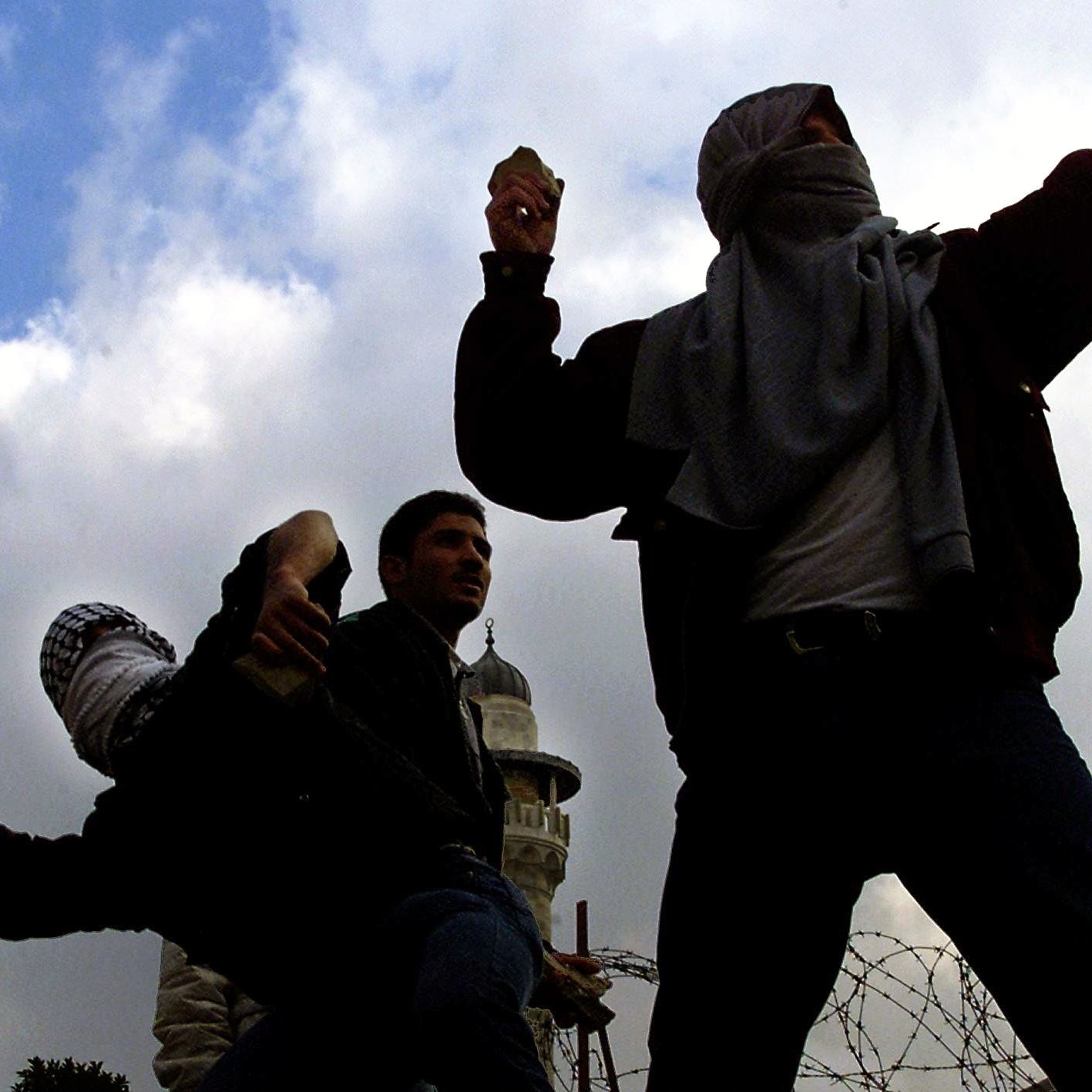 L'image montre plusieurs personnes en train de lancer des projectiles, probablement dans un contexte de confrontation ou de manifestation. Ils portent des vêtements sombres et ont le visage partiellement couvert, ce qui suggère qu'ils cherchent à dissimuler leur identité. En arrière-plan, on aperçoit une tour, probablement une mosquée, et des barbelés, renforçant l'idée d'un environnement tendu ou conflictuel. Le ciel est nuageux, ce qui contribue à l'atmosphère dramatique de la scène.