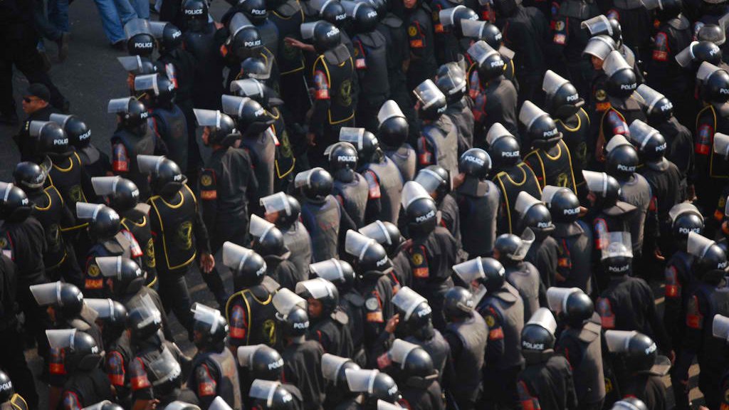 L'image montre un grand groupe de policiers en uniforme, portant des casques et des protections. Ils sont disposés de manière organisée et semblent être en formation, probablement lors d'une manifestation ou d'un événement de sécurité. Les uniformes sont généralement noirs avec des éléments de couleur, et on peut voir des panneaux de protection en plastique. L'atmosphère semble intense et sérieuse, reflétant une situation de gestion de la foule.
