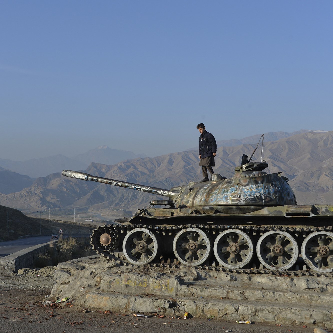 في الصورة، يظهر رجل واقفًا على دبابة قديمة متوقفة بجانب طريق. الدبابة تحمل آثار الزمن، حيث أن لونها باهت ومغطى ببعض الصدأ. في الخلفية، يمكن رؤية الجبال ذات الألوان الترابية، مما يضفي طابعًا طبيعيًا على المشهد. الطريق الذي يمر بجوار الدبابة يظهر خاليًا تقريبًا، مع وجود سيارة تسير في اتجاه معين. السماء صافية وضياء الشمس يضفي شعورًا بالهدوء على الصورة.