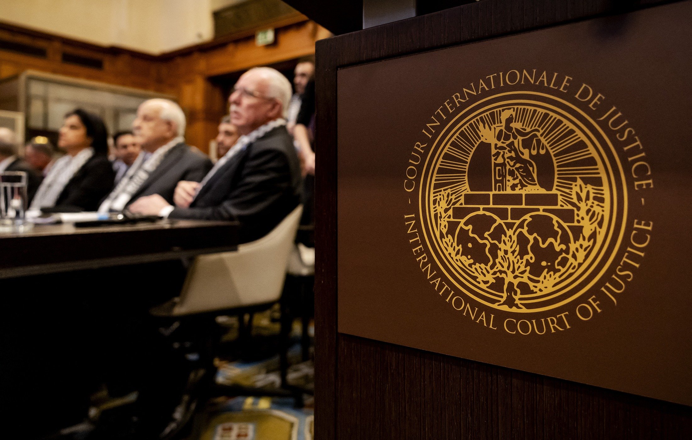 L'image montre une scène à la Cour internationale de justice. Au premier plan, on peut voir le symbole de la Cour, qui est un emblème distinctif. En arrière-plan, plusieurs personnes sont assises, probablement des membres du tribunal ou des délégués, attendant une session ou un discours. L'environnement semble formel, avec une décoration typique d'une institution judiciaire.