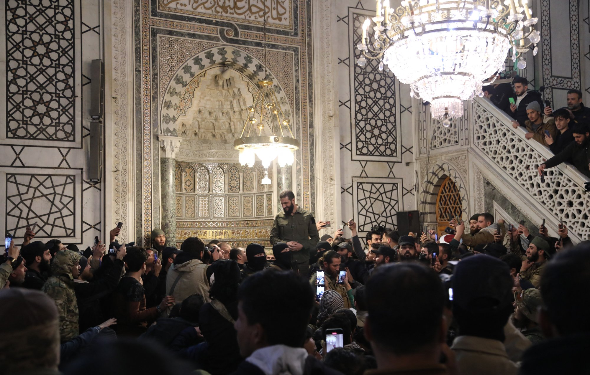 تظهر الصورة مجموعة من الأشخاص في مكان داخلي مزين بتفاصيل معمارية جميلة. هناك شخص يقف في الوسط، يبدو أنه يتحدث أو يلقي كلمة، بينما يحيط به حشد من المشاركين الذين يلتقطون الصور بأجهزتهم المحمولة. الإضاءة في المكان تتضمن ثريا كبيرة تعكس طابعًا تاريخيًا وثقافيًا، مع وجود عناصر زخرفية معمارية مميزة على الجدران. يبدو أن الحدث مهم وجذب انتباه الحضور بشكل كبير.