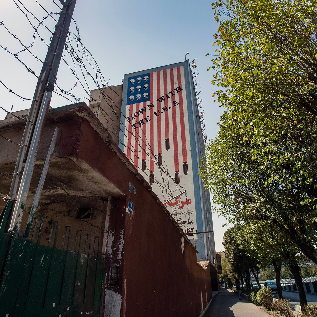 تصویر نشان‌دهنده دیواری است که بر روی آن پرچم آمریکا نقاشی شده است. این پرچم به گونه‌ای طراحی شده که بخشی از آن به صورت معکوس و غیرمتعارف نمایش داده شده است. در پس‌زمینه، fences (حصارهایی) با میله‌های تیز دیده می‌شود. همچنین درختان و یک پیاده‌رو در کنار دیوار وجود دارد. نور آفتاب به تصویر زیبایی افزوده است و فضا به طور کلی حس خاصی از تاریخ و سیاست را منتقل می‌کند.