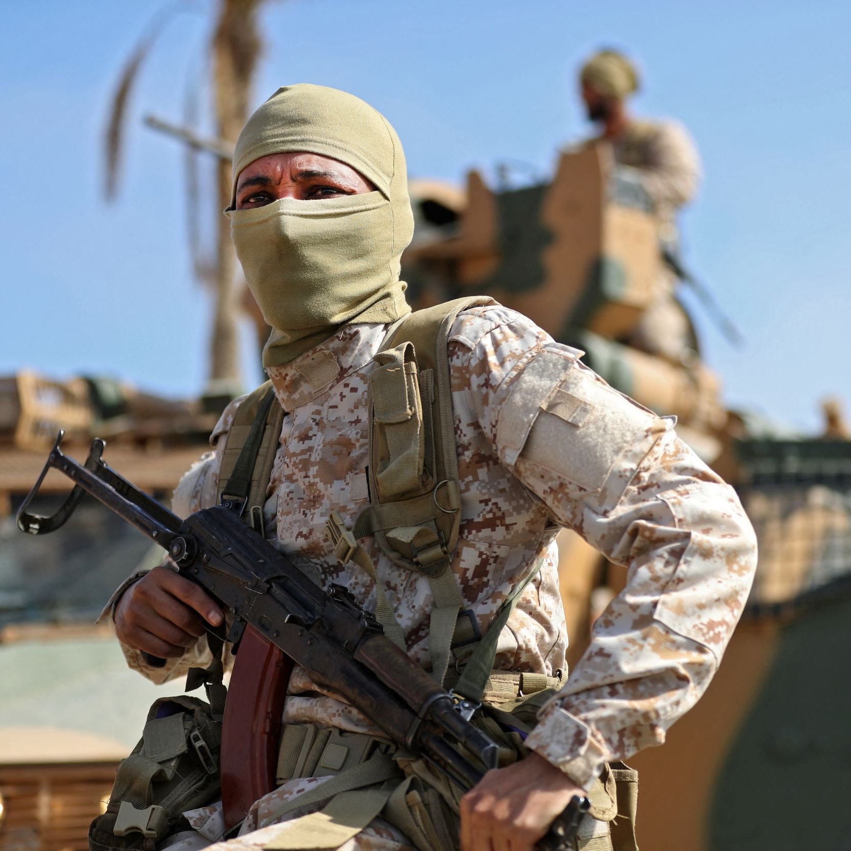 L'image montre un soldat portant un uniforme camouflé et un masque couvrant le bas de son visage. Il est équipé d'une arme et se tient en position vigilante, probablement en mission. En arrière-plan, on peut apercevoir un véhicule militaire et un autre soldat. Le ciel est dégagé, suggérant une ambiance ensoleillée et une atmosphère de tension.
