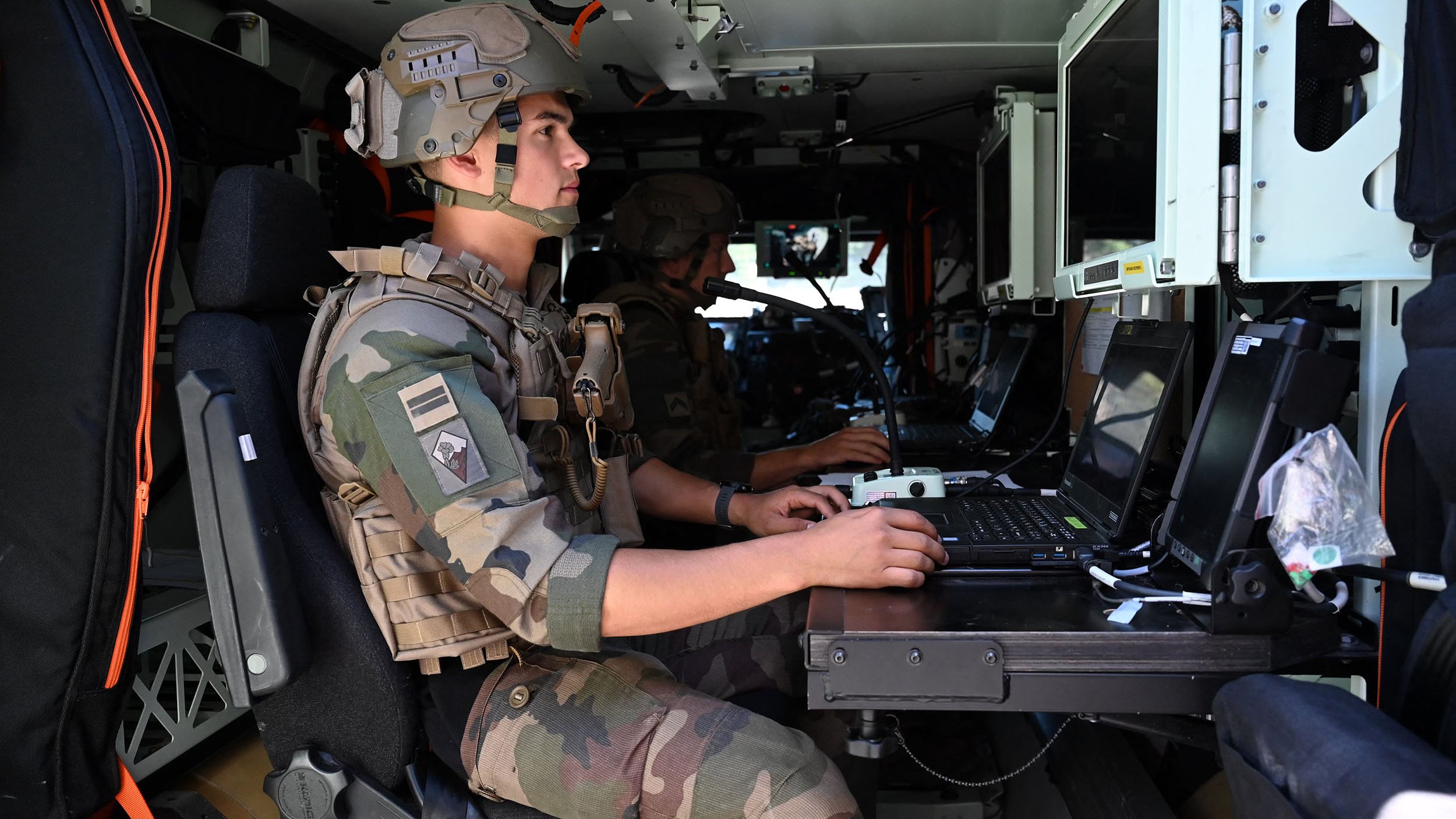 L'image montre deux soldats à l'intérieur d'un véhicule militaire. Ils portent des uniformes camouflés et des casques. L'un des soldats est assis devant un ordinateur portable, concentré sur son travail. L'intérieur du véhicule est équipé de plusieurs écrans et dispositifs technologiques. L'atmosphère semble sérieuse, indiquant une mission ou une opération en cours.