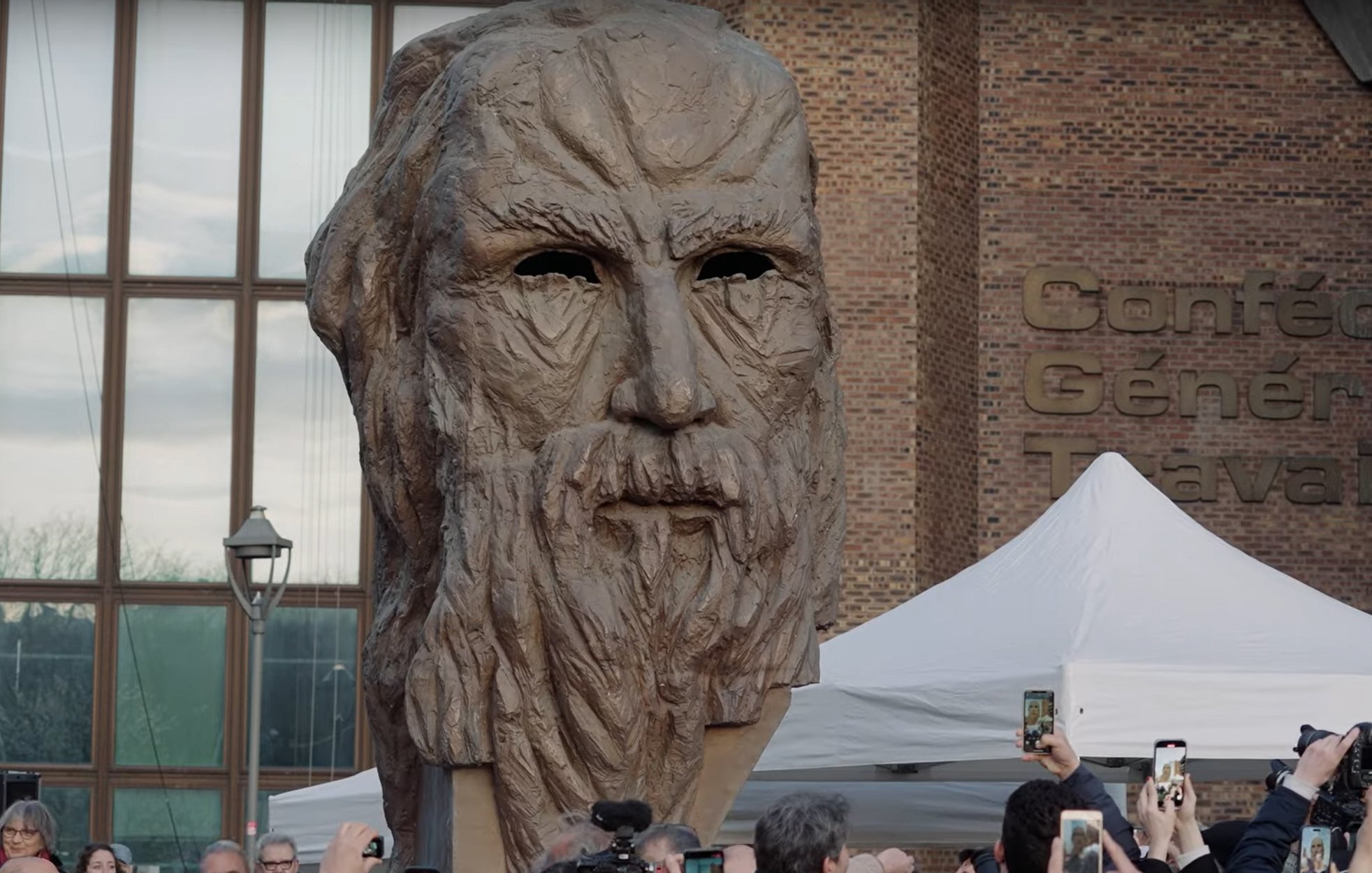 L'image montre une grande sculpture représentant un visage ancien, probablement celui d'une figure historique ou mythologique, avec des traits marqués et une barbe. Elle est entourée d'une foule de personnes qui semblent prendre des photos avec leurs téléphones. Il y a aussi des tentes et un bâtiment visible en arrière-plan, ce qui indique qu'il s'agit probablement d'un événement public ou d'une exposition. L'atmosphère semble être festive et attentive, centrée sur l'œuvre sculpturale.
