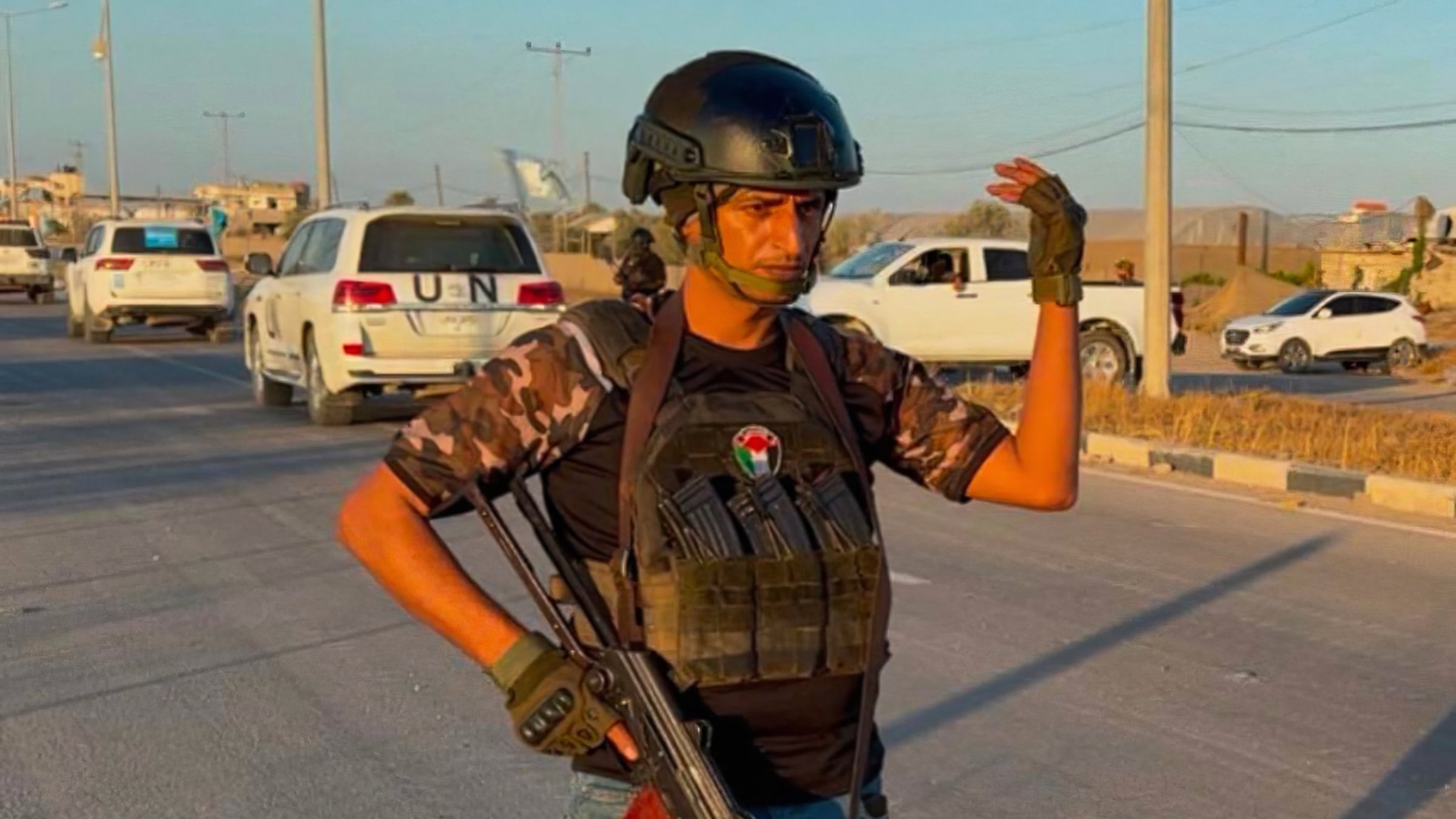 Un homme armé, en tenue militaire, se tient sur une route avec des voitures en arrière-plan.