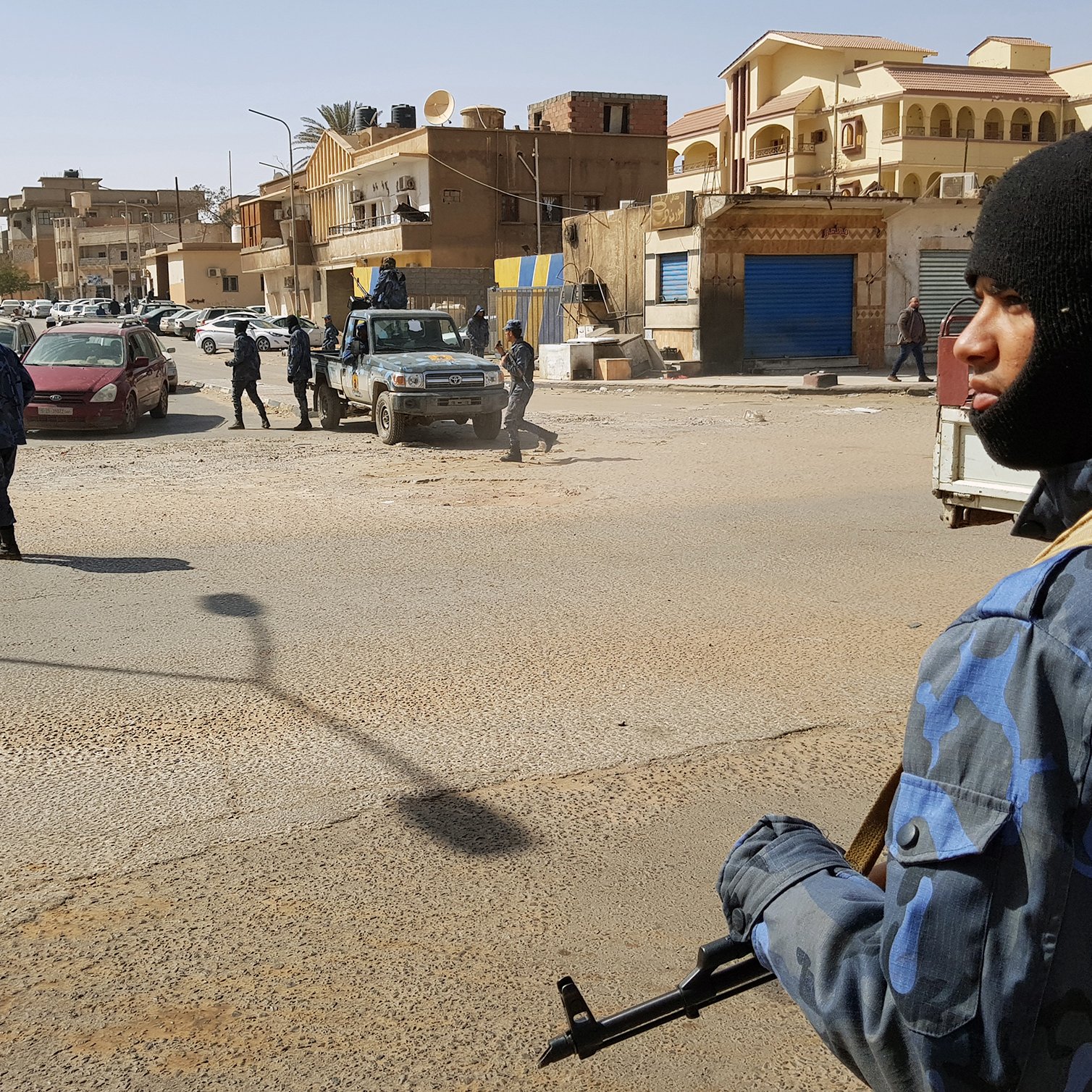 The image depicts a street scene in an urban environment where armed security personnel are present. One individual is prominently featured in the foreground, wearing a dark mask and tactical gear, holding a firearm. In the background, other security personnel are visible, some moving and others standing near vehicles, including a jeep. The street appears to be busy, with buildings lining either side and several parked cars. The setting suggests heightened security or law enforcement activity.