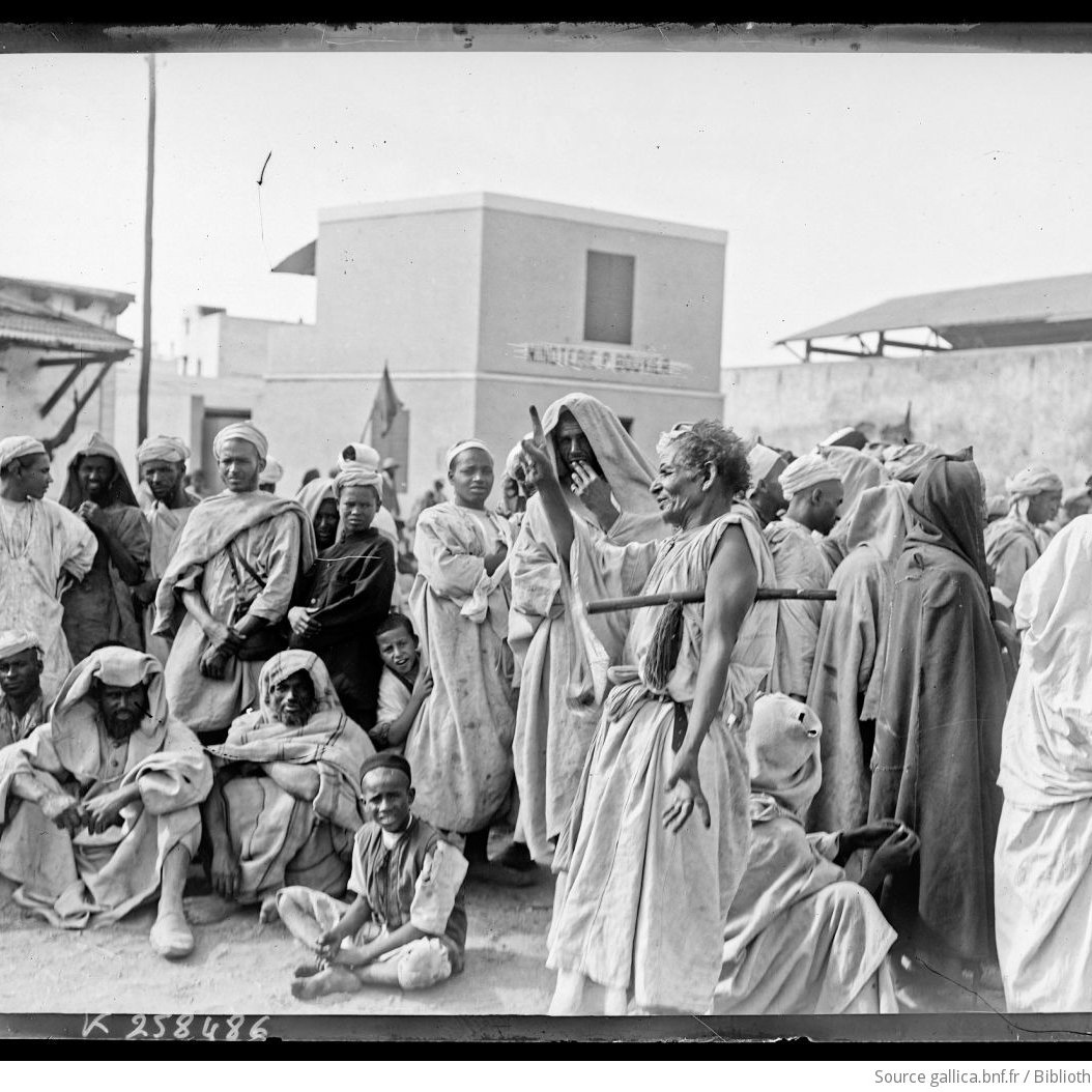 L'image montre un groupe de personnes rassemblées dans un environnement extérieur. Elles portent des vêtements traditionnels et semblent discuter ou observer quelque chose au centre. L'atmosphère est marquée par une certaine sérénité, mais aussi par une curiosité collective. En arrière-plan, on peut distinguer des bâtiments, suggérant un lieu structuré, potentiellement un marché ou un rassemblement communautaire. La lumière naturelle éclaire la scène, ajoutant à l'authenticité de l'ambiance.
