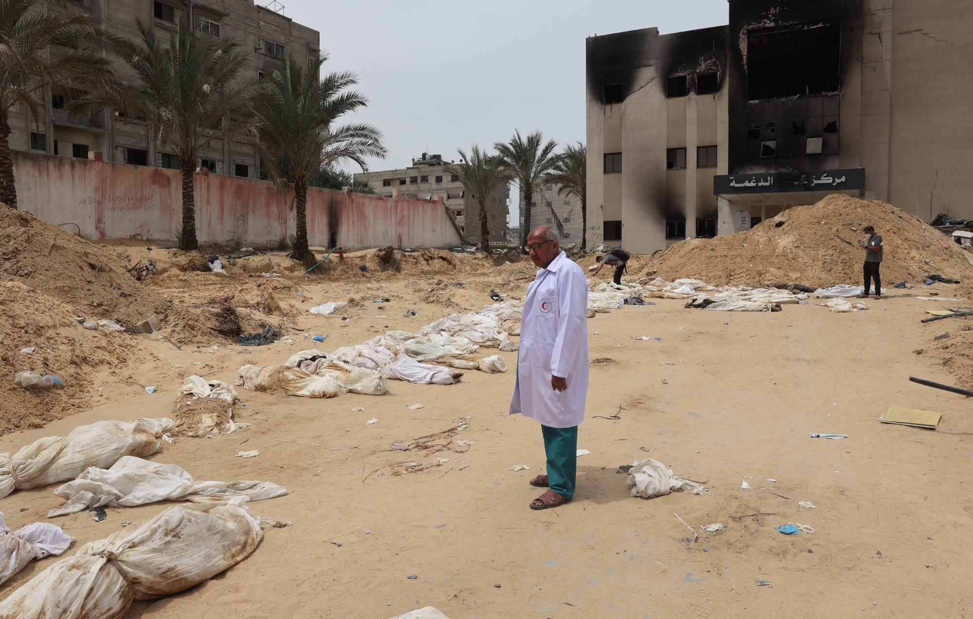 L'image montre un homme en blouse blanche se tenant debout sur un terrain désertique, entouré de sacs ressemblant à des corps. En arrière-plan, on peut voir des bâtiments endommagés et des morceaux de débris éparpillés sur le sol. La scène évoque un environnement de désastre, avec une atmosphère sombre et préoccupante, suggérant des événements tragiques. Des palmiers se dressent également sur le site, ajoutant un contraste avec la gravité de la situation.