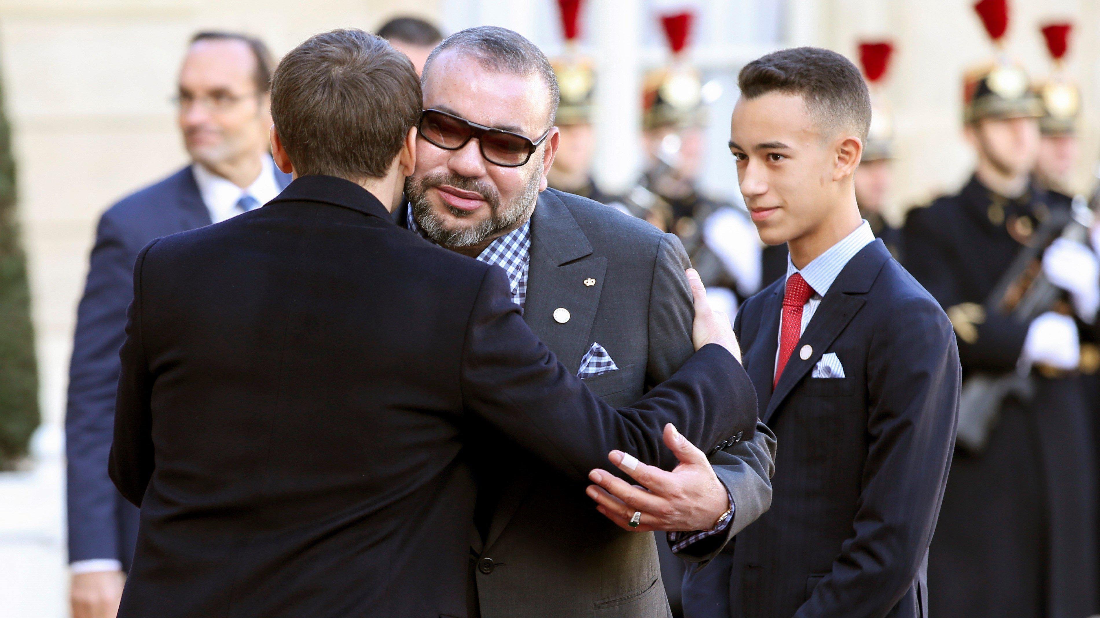 L'image montre une rencontre officielle entre deux hommes. L'un des hommes, vêtu d'un costume élégant, semble accueillir l'autre avec une accolade amicale. L'autre homme, portant des lunettes de soleil et un costume gris, se tient à côté d'un jeune garçon en costume sombre et cravate. En arrière-plan, on aperçoit des gardes en uniforme, ce qui indique un contexte cérémonial. L'atmosphère semble conviviale et respectueuse.