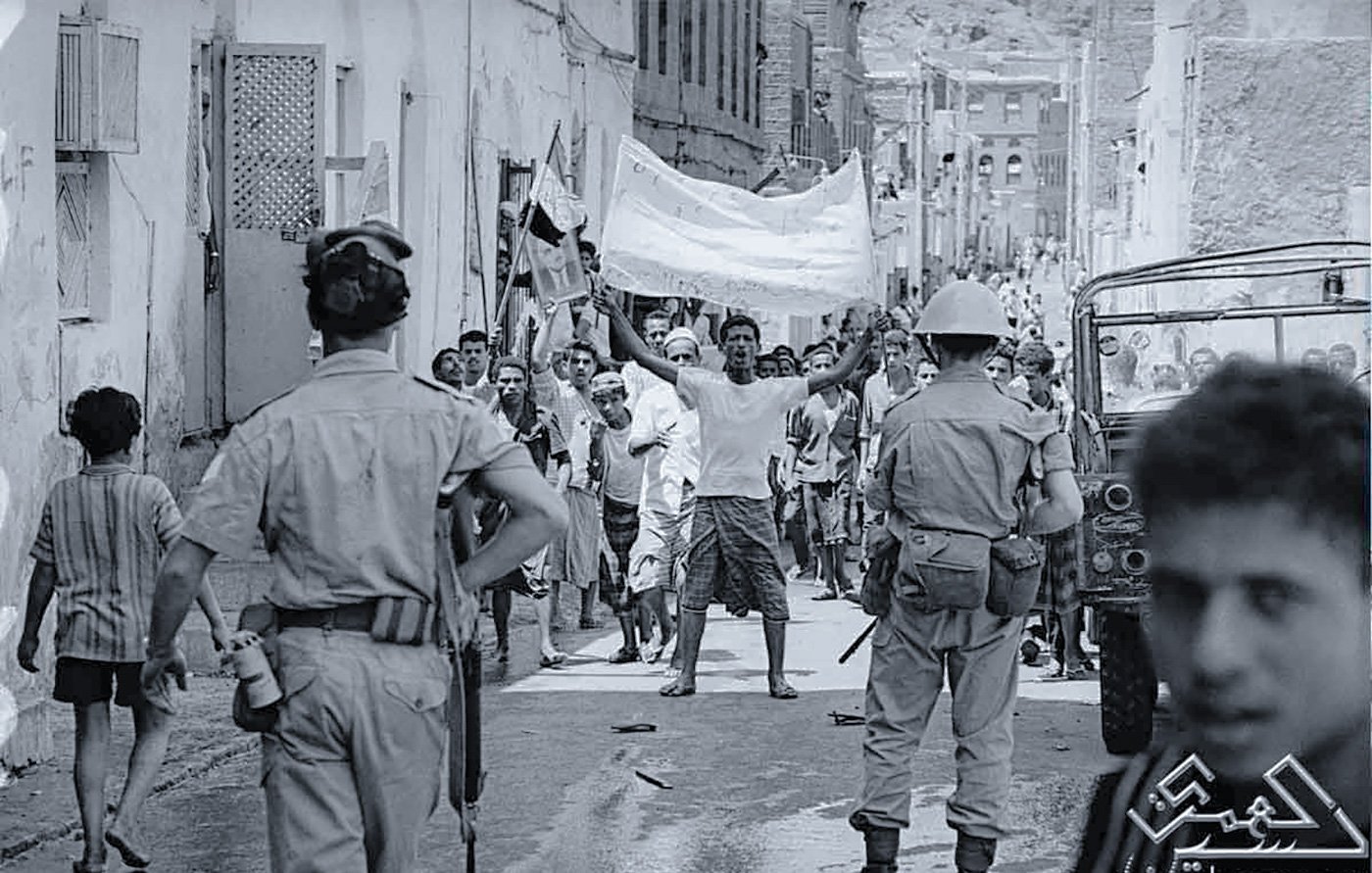 تظهر الصورة مجموعة من الأشخاص في شارع ضيق، يبدو أنهم في خضم حدث أو تظاهرة. في المقدمة، هناك رجل يرفع يديه حاملاً لافتة، بينما يتواجد أفراد من قوات الأمن بالقرب منه. يمكن رؤية الأطفال والبالغين في الخلفية، بعضهم يبدو مهتماً بالحدث. الملابس والإعداد تعكس فترة زمنية معينة، كما يظهر في التفاصيل المعمارية للمنازل. المشهد يحتوي على أجواء من الحماس والتوتر.