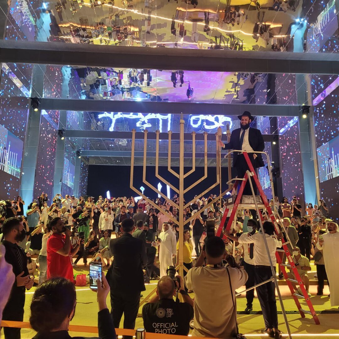 الصورة تظهر حدثًا احتفاليًا في مكان كبير. في المركز، يوجد هيكل على شكل منارة (المناورة)، مما يشير إلى الاحتفال بمناسبة معينة. يتم تنظيم الحضور حول الهيكل، وهناك أشخاص يحملون هواتفهم لتصوير الحدث. بعض الحضور يرتدون ملابس تقليدية، بينما يرتدي آخرون ملابس رسمية. هناك إضاءة ملونة وتفاصيل زينة جميلة في الأجواء، مما يضيف جوًا احتفاليًا حيويًا.