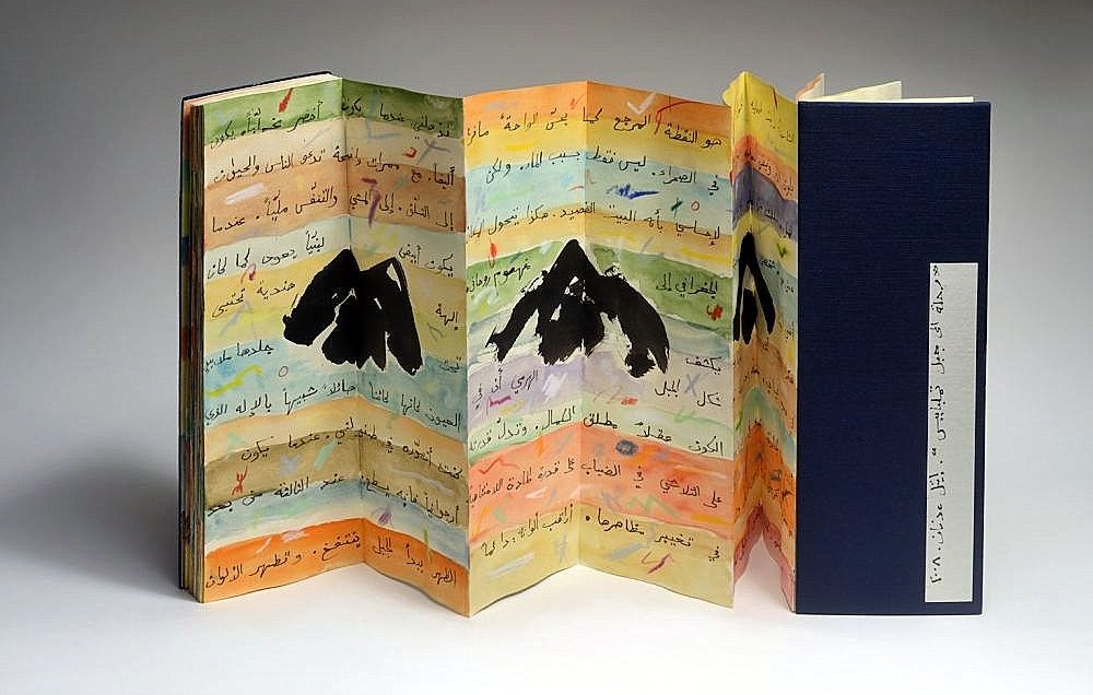 L'image montre un livre artistique aux pages colorées, présentant des illustrations de montagnes. Les pages sont pliées en accordéon et comportent des inscriptions en arabe. La palette de couleurs est variée, avec des nuances de jaune, bleu et orange, ajoutant une dimension vibrante à l'ensemble. Le livre est partiellement ouvert, révélant sa structure unique et sa conception créative. Une couverture noire est visible à droite, contrastant avec les couleurs vives des pages intérieures.