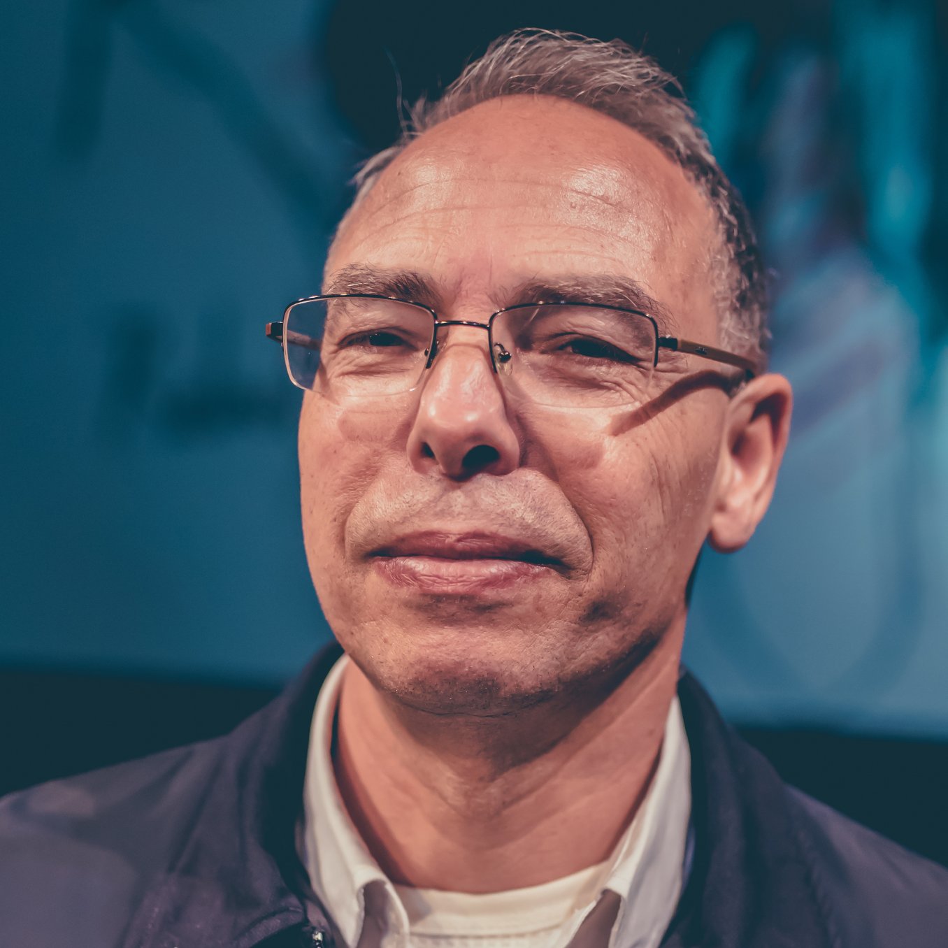 Homme souriant avec des lunettes, cheveux courts et gris, fond flou en arrière-plan.