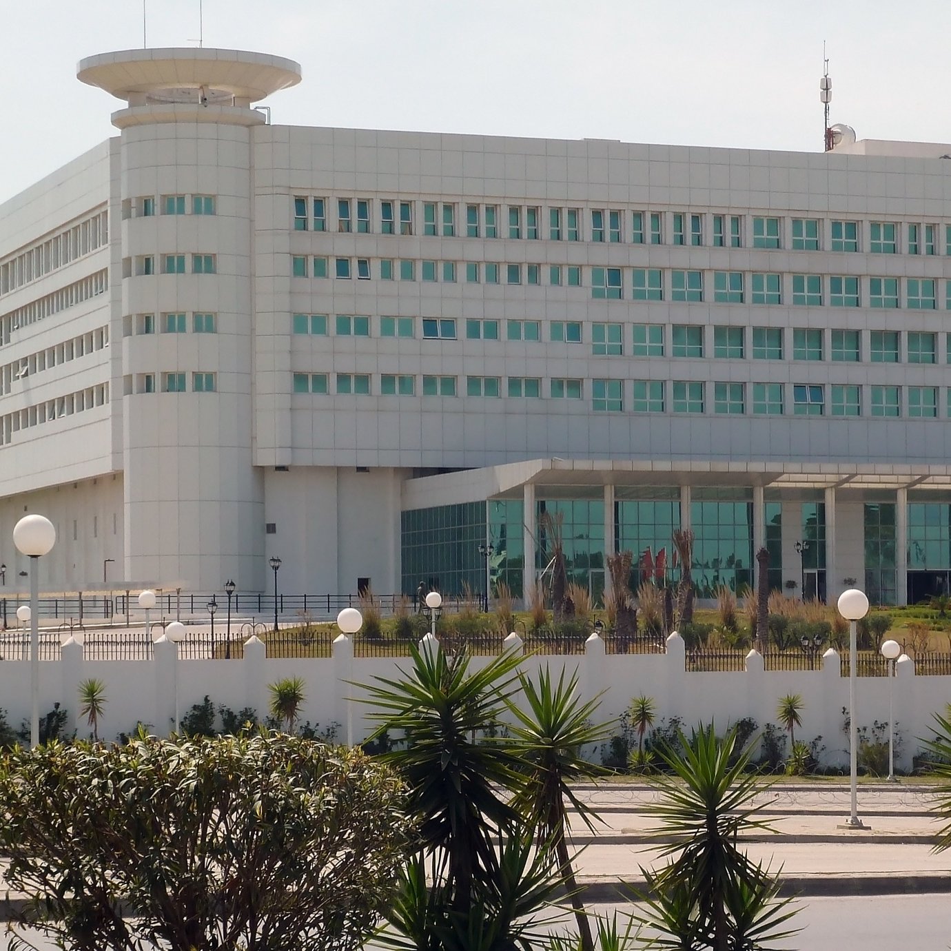 L'image montre un bâtiment moderne avec une architecture contemporaine. Il est de couleur blanche, avec de grandes fenêtres et un design carré. À l'avant, il y a une entrée spacieuse avec des colonnes et des sculptures. Le paysage environnant est bien entretenu, avec des plantes et des lampadaires. On peut également voir un drapeau en haut du bâtiment, ce qui indique qu'il pourrait s'agir d'un bâtiment gouvernemental ou institutionnel.