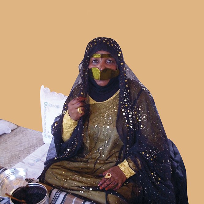 L'image montre une femme vêtue d'une tenue traditionnelle ornée de motifs et de broderies. Elle porte un voile noir qui couvre son visage en grande partie, laissant seulement ses yeux visibles, ce qui ajoute une touche de mystère. Sa robe est richement décorée, avec des couleurs sombres et des embellissements scintillants. À côté d'elle, on aperçoit des objets traditionnels, ce qui suggère une ambiance culturelle. L'arrière-plan est d'une couleur unie, mettant en valeur sa présence.