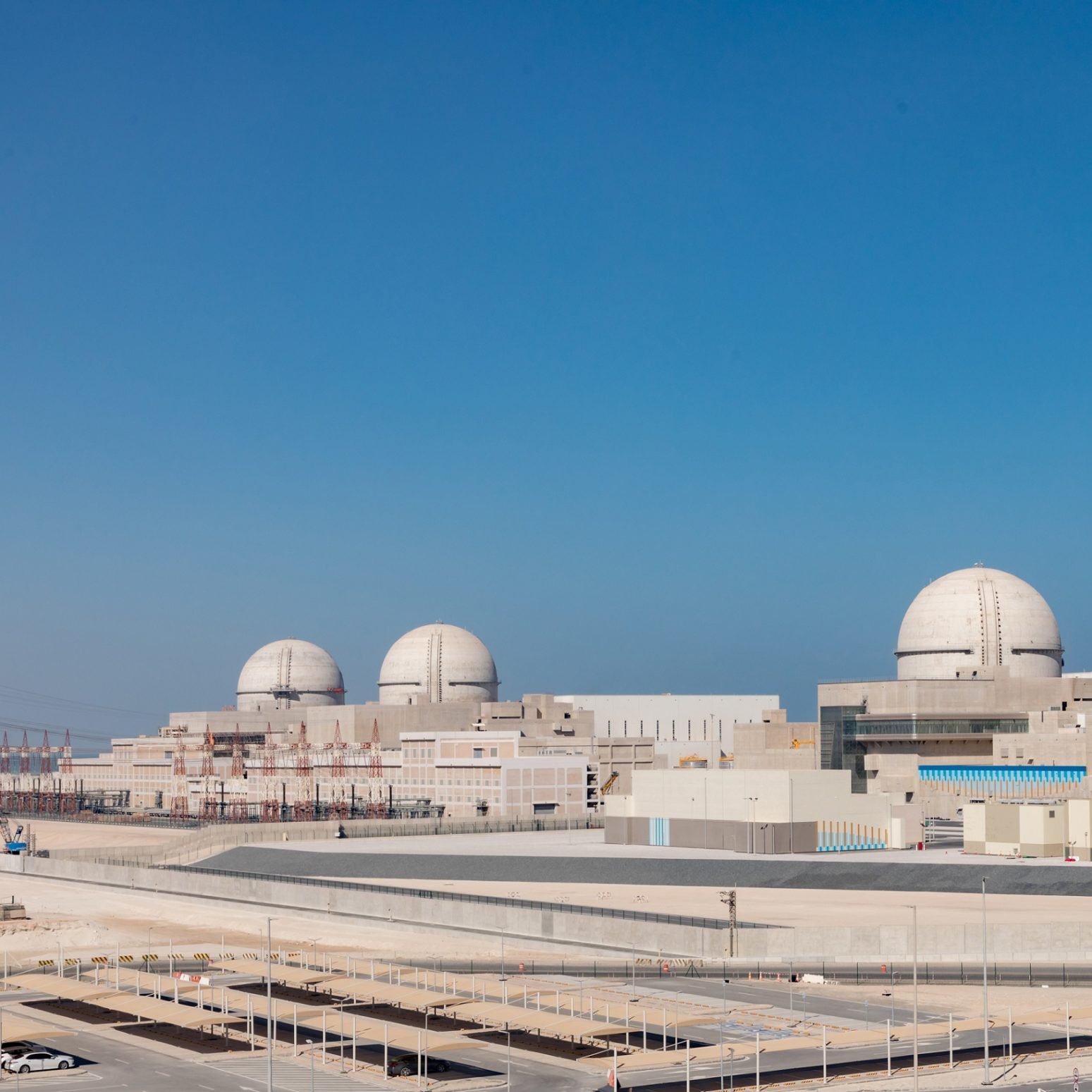 L'immagine mostra una centrale nucleare moderna, caratterizzata da tre imponenti cupole bianche che emergono dal complesso. La struttura presenta un design industriale, con vari edifici di colore chiaro che si estendono in un'area ampia e ben organizzata. Il cielo è sereno e blu, suggerendo una bella giornata. In primo piano, si può notare un'area ben curata e delle strade di accesso, tipiche delle installazioni di questo tipo.