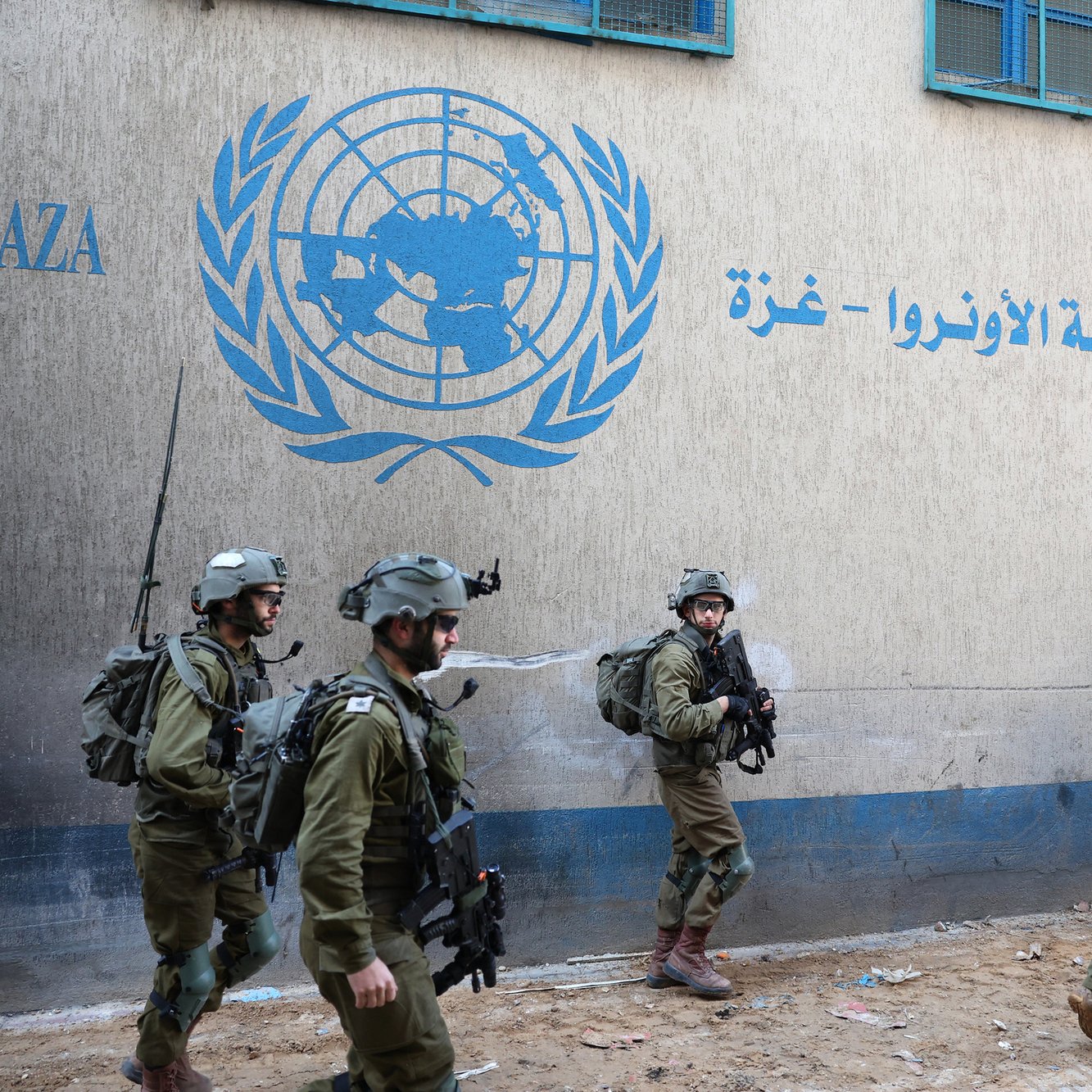 L'immagine mostra un gruppo di soldati in uniforme militare che camminano davanti a un edificio con un logo delle Nazioni Unite, situato nella regione di Gaza. I soldati indossano elmetti e sono equipaggiati per situazioni di operazioni di sicurezza. L'atmosfera è seria e il contesto suggerisce una situazione di tensione o di operazioni militari nella zona. Sul muro dell'edificio si vedono scritte in arabo e in inglese, che indicano che si tratta di un ufficio dell'UNRWA, l'Agenzia delle Nazioni Unite per i Rifugiati Palestinesi.