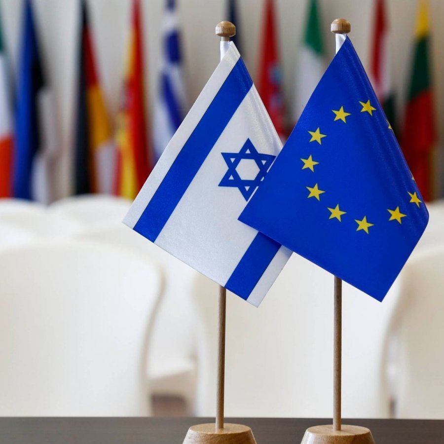 تظهر الصورة علمين صغيرين مثبَّتين على قاعدة خشبية. العلم الأول هو علم إسرائيل، الذي يحتوي على نجمتين زرقاوين على خلفية بيضاء وبينهما خطان أفقيان زرقاوان. العلم الثاني هو علم الاتحاد الأوروبي، الذي يحتوي على دائرة من دوائر النجوم الذهبية على خلفية زرقاء. خلفهم، يمكن رؤية مجموعة من الأعلام المختلفة المعلقَة، مما يوحي بأجواء تتعلق بالفعاليات الدولية أو الاجتماعات.